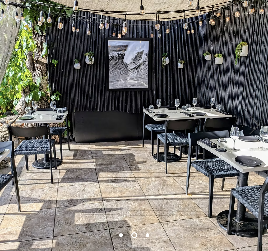 An outdoor dining area with black tables and chairs, set with wine glasses and black plates. A black wall decorated with small white planters holding green plants is in the background, topped with string lights and a large framed ocean wave photograph. There are trees and greenery visible on the left side.