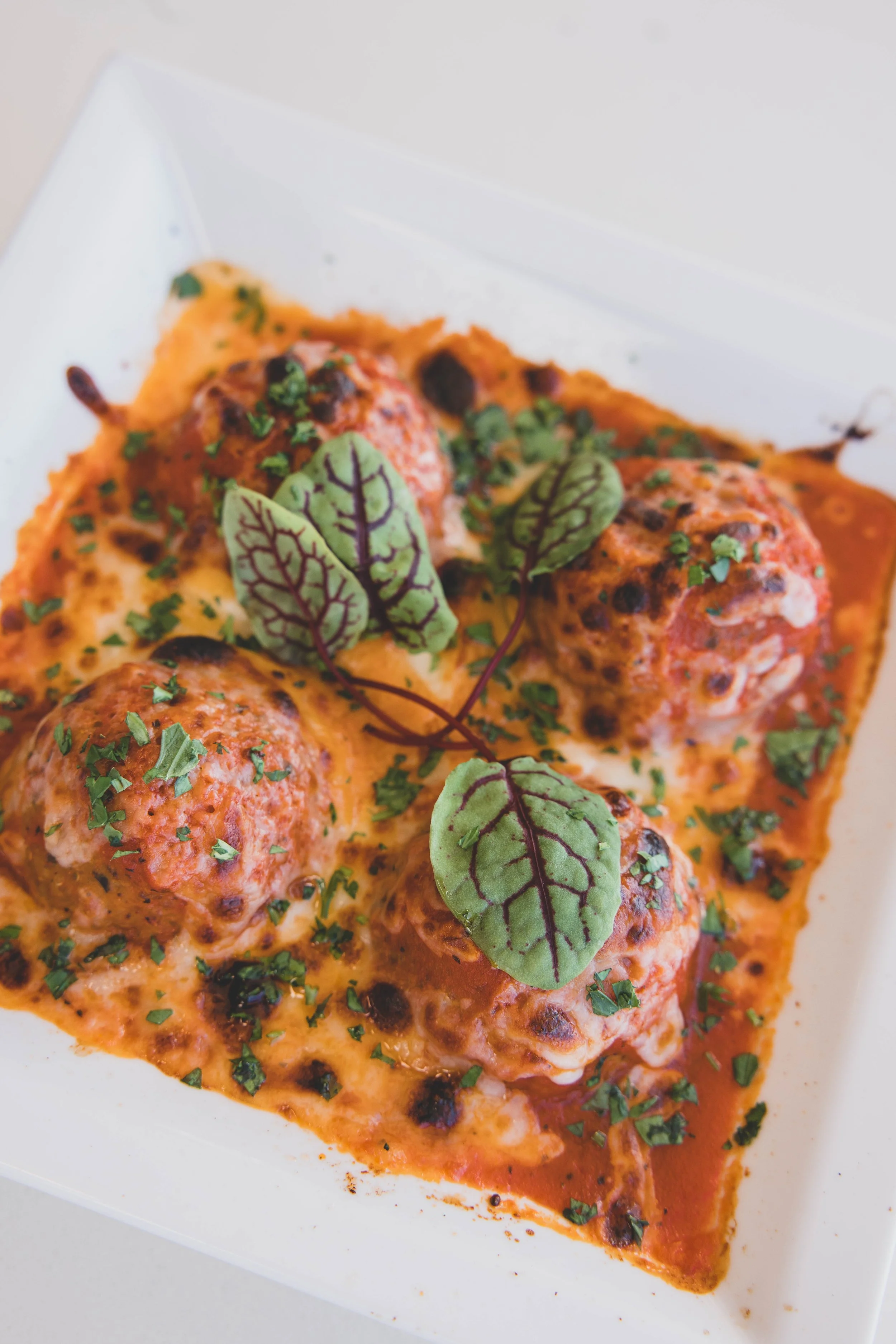 Close-up of baked meatballs in tomato sauce, garnished with chopped herbs and fresh basil leaves, served in a white rectangular dish.