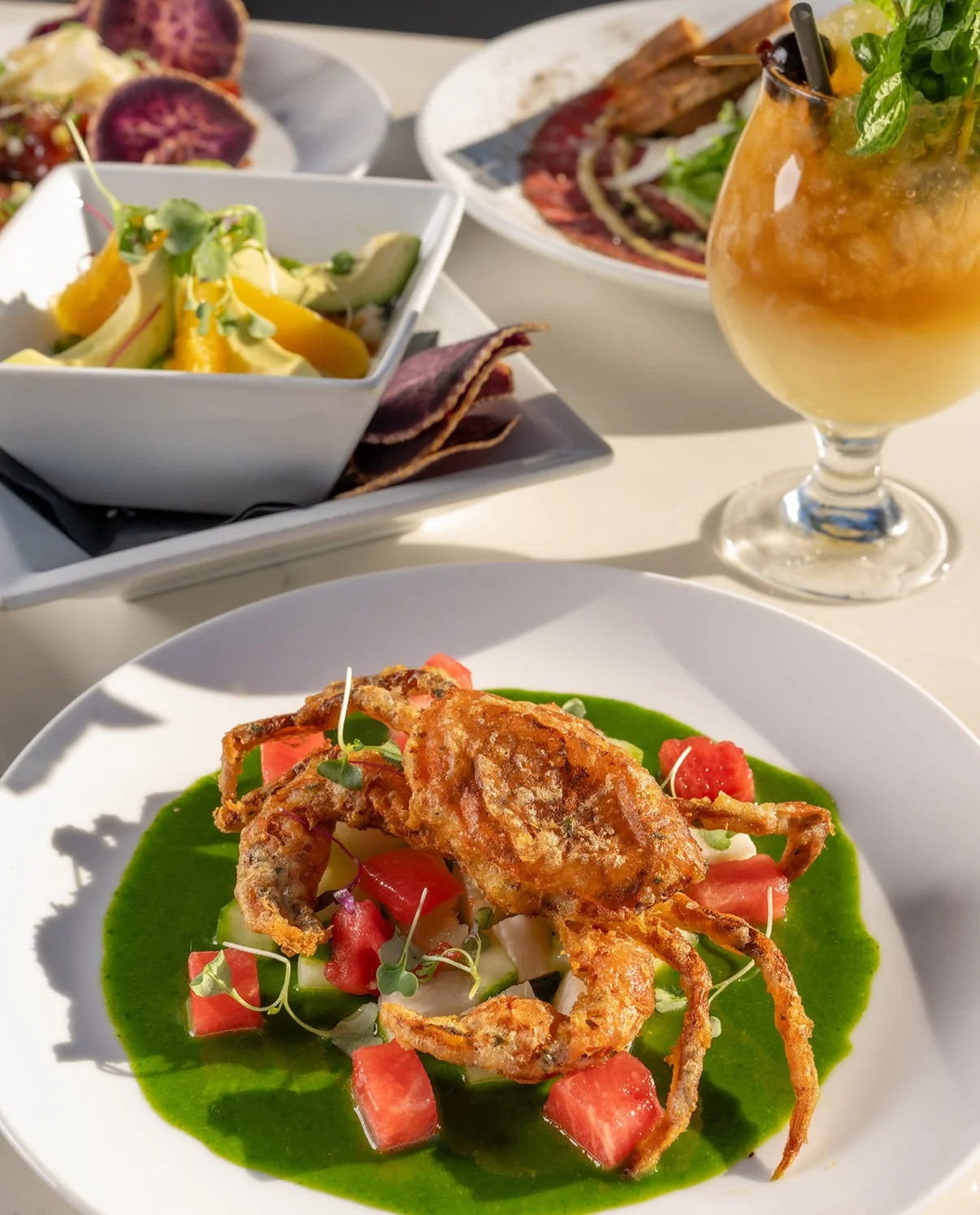 Fried calamari served on a white plate with green sauce, garnished with diced tomatoes, onions, and microgreens, with a grassy herb drink and other dishes in the background.