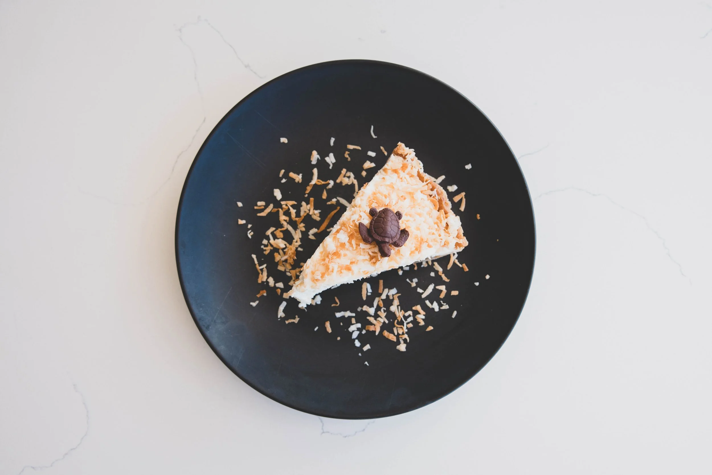Slice of coconut cake garnished with a turtle-shaped chocolate piece on a black plate, with coconut shavings around it, on a white surface.