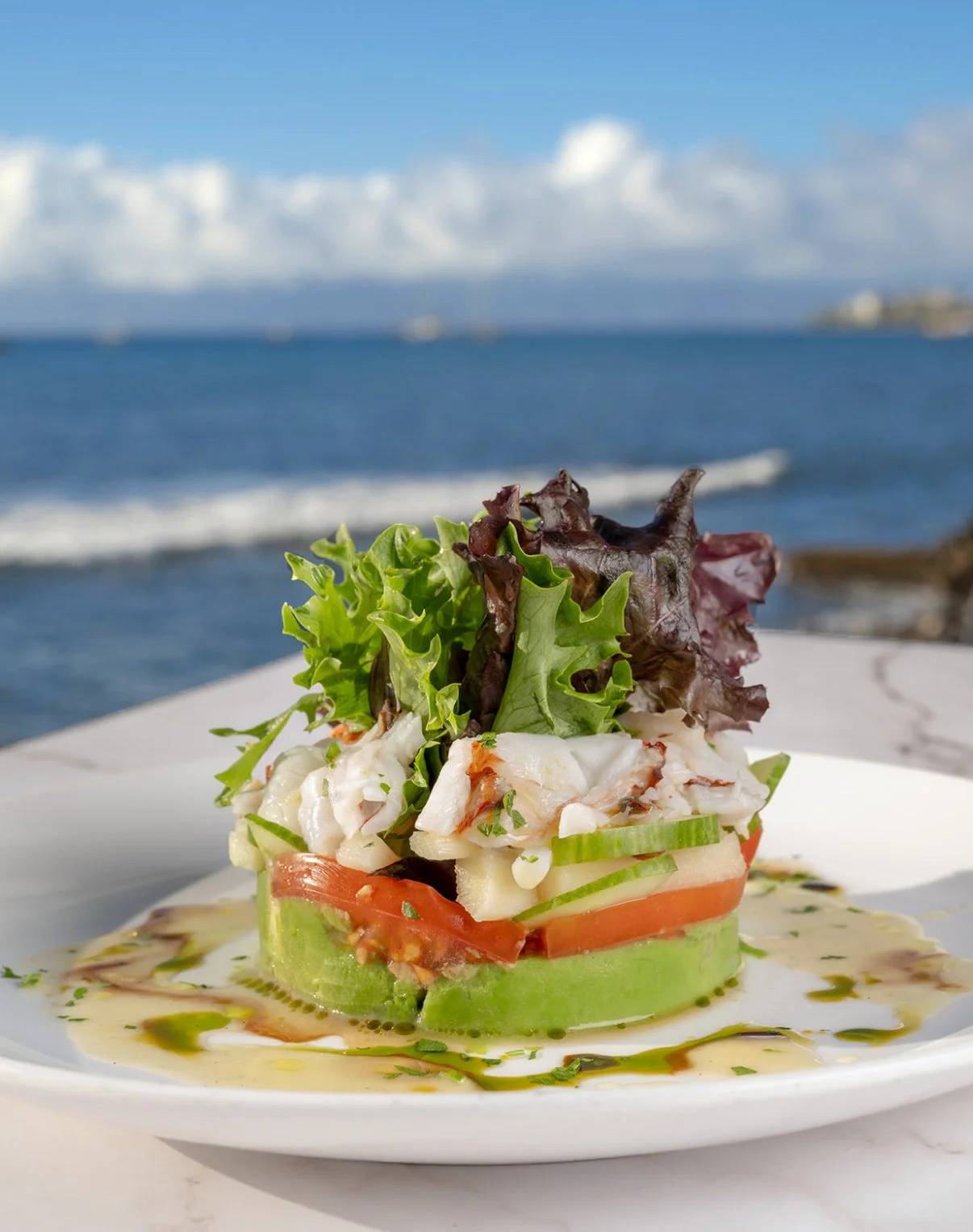 Seafood salad with lettuce, tomato, cucumber on a round slice of avocado, drizzled with vinaigrette on a white plate at the beach.
