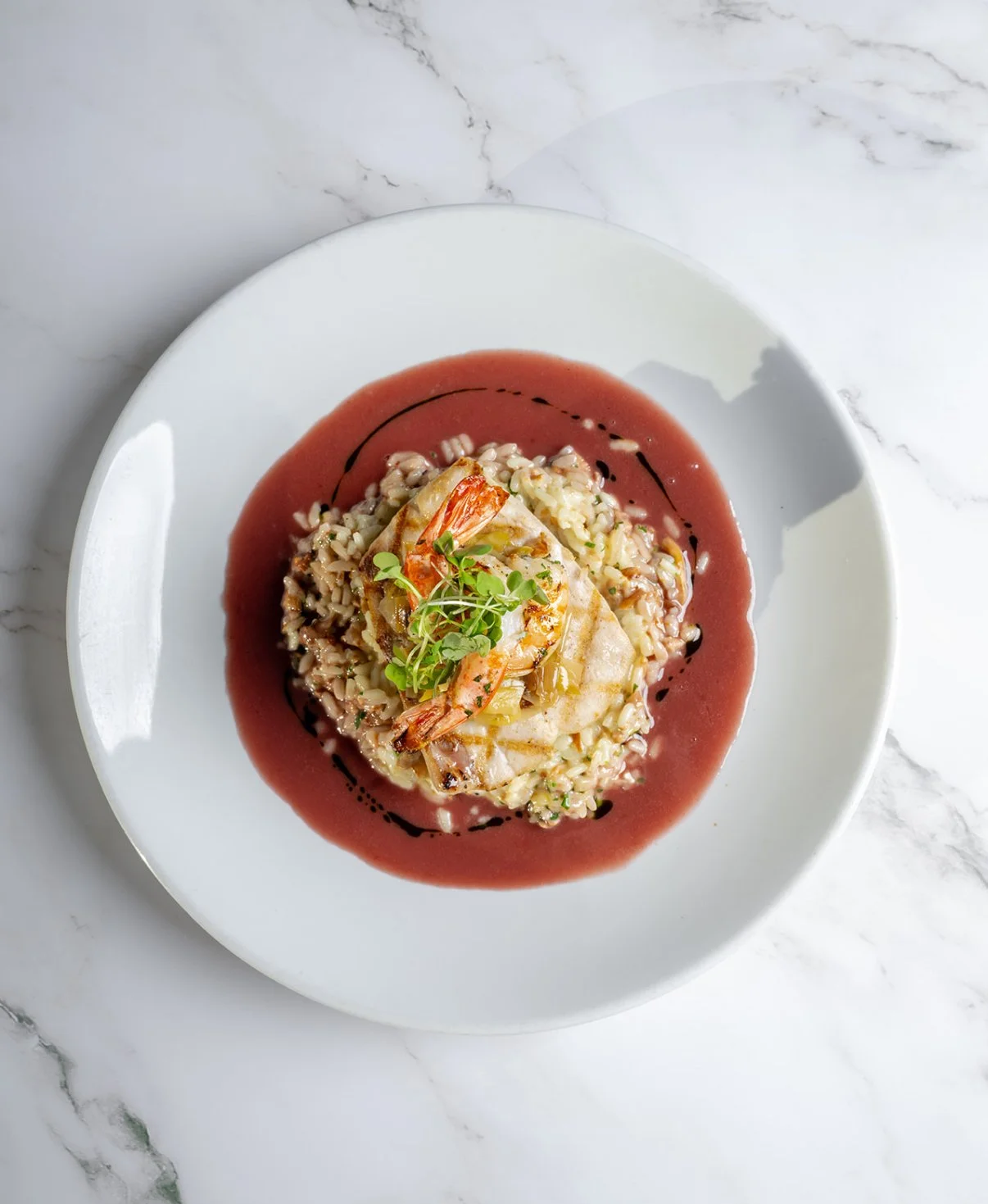 A white plate with a seafood risotto topped with shrimp, garnished with herbs, and served with a red sauce and a drizzle of balsamic reduction, on a marble surface.