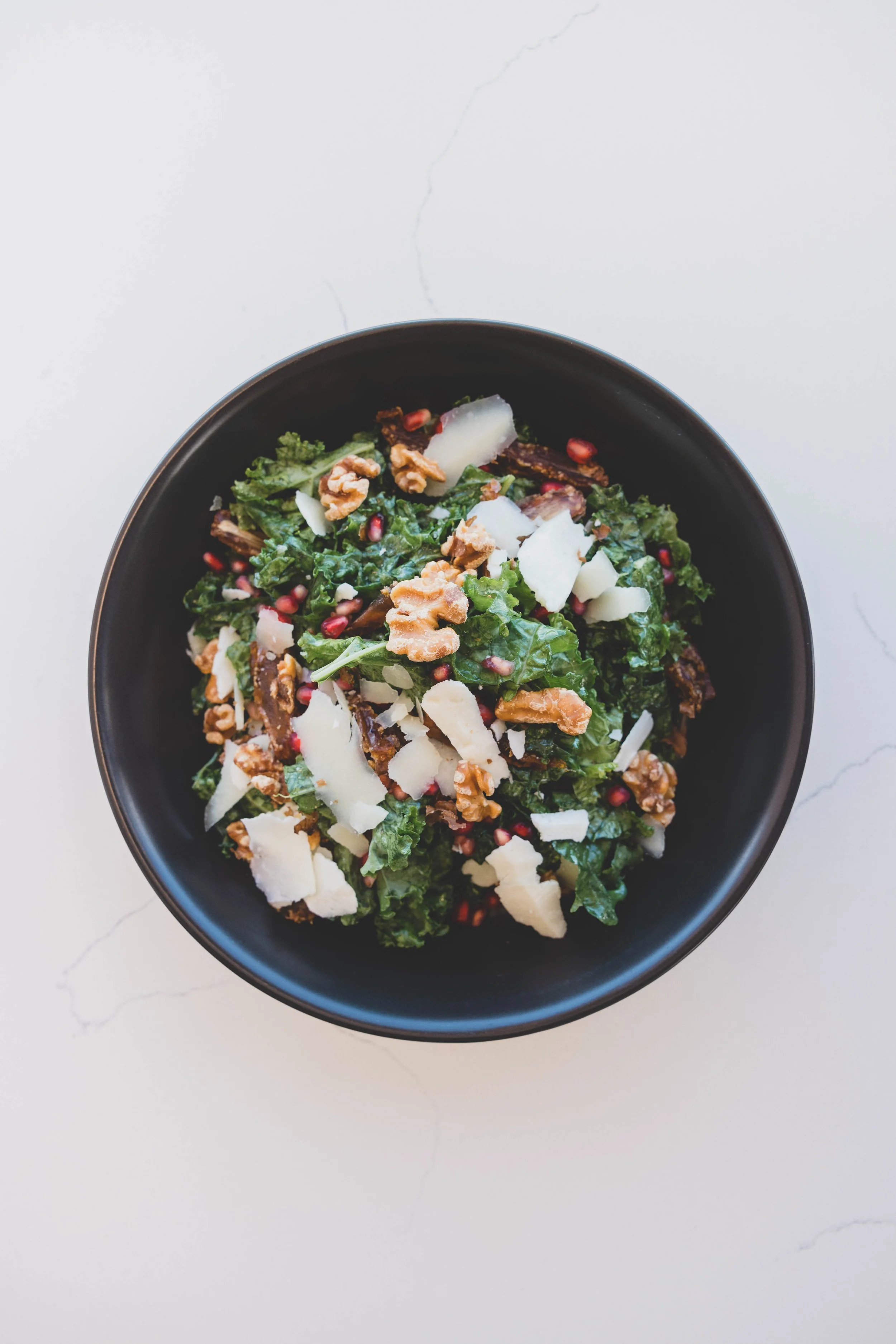 A bowl of kale salad topped with walnuts, cheese shavings, pomegranate seeds, and possibly dried cranberries on a white surface.