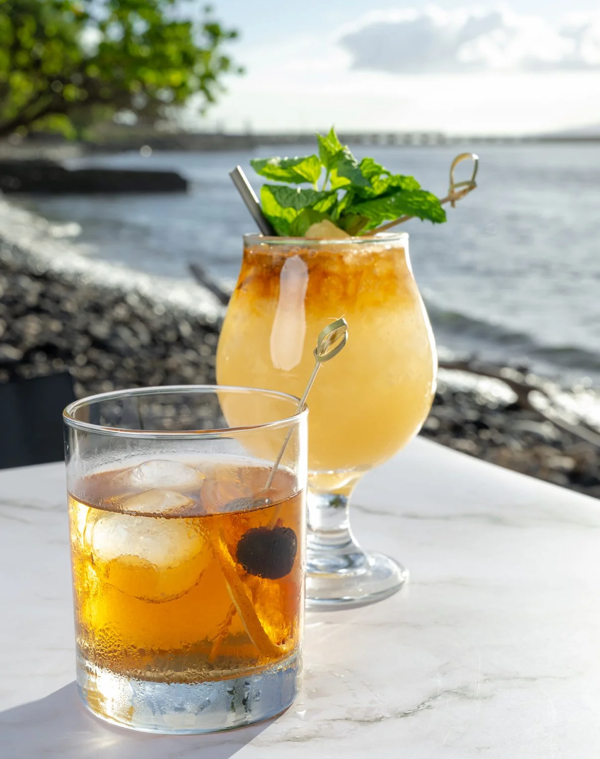 Two cocktails on a white marble table with a lakeshore in the background. One drink is a glass of dark liquid with ice and a cherry garnish, the other is a yellowish tropical drink garnished with mint leaves.