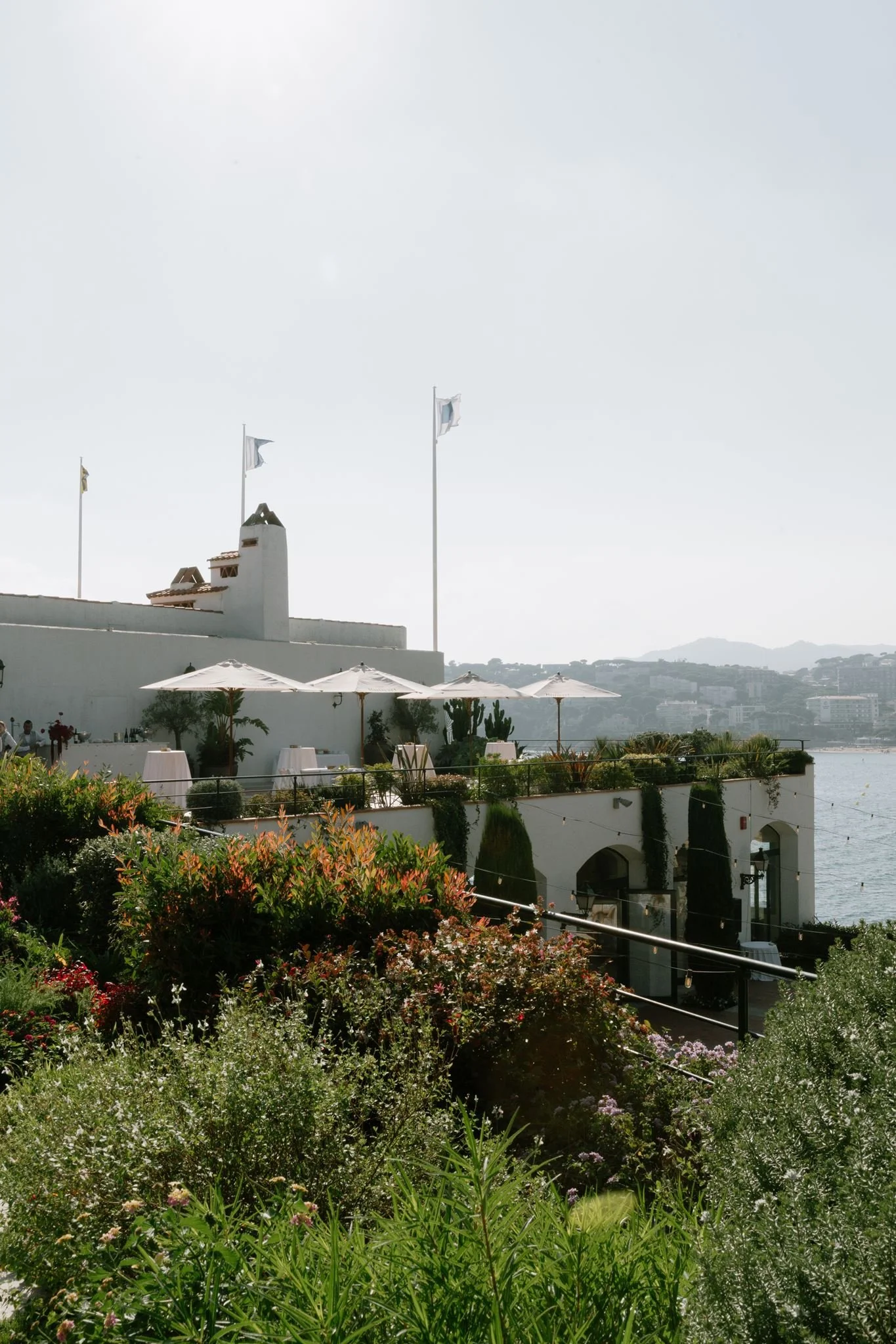 A white building with a chimney and flags flying on poles, surrounded by greenery and flowers, with hills in the background.