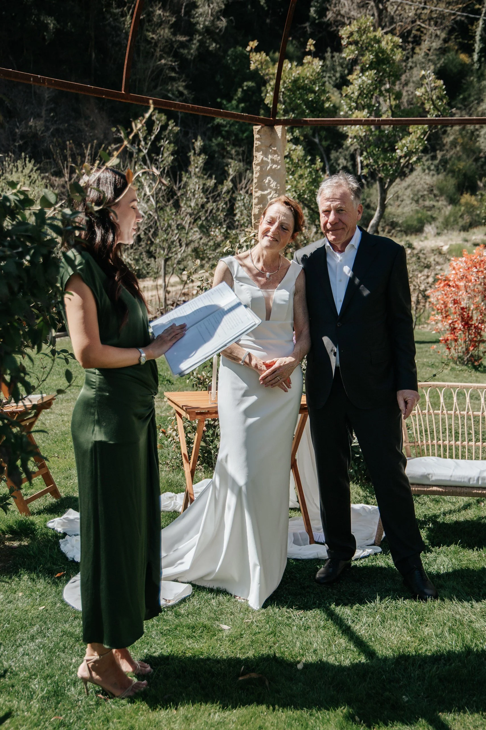 A wedding ceremony outdoors with a bride and groom standing close together, a woman officiant reading from a book, and a woman in a green dress standing nearby. The setting is lush and green with trees and shrubs.