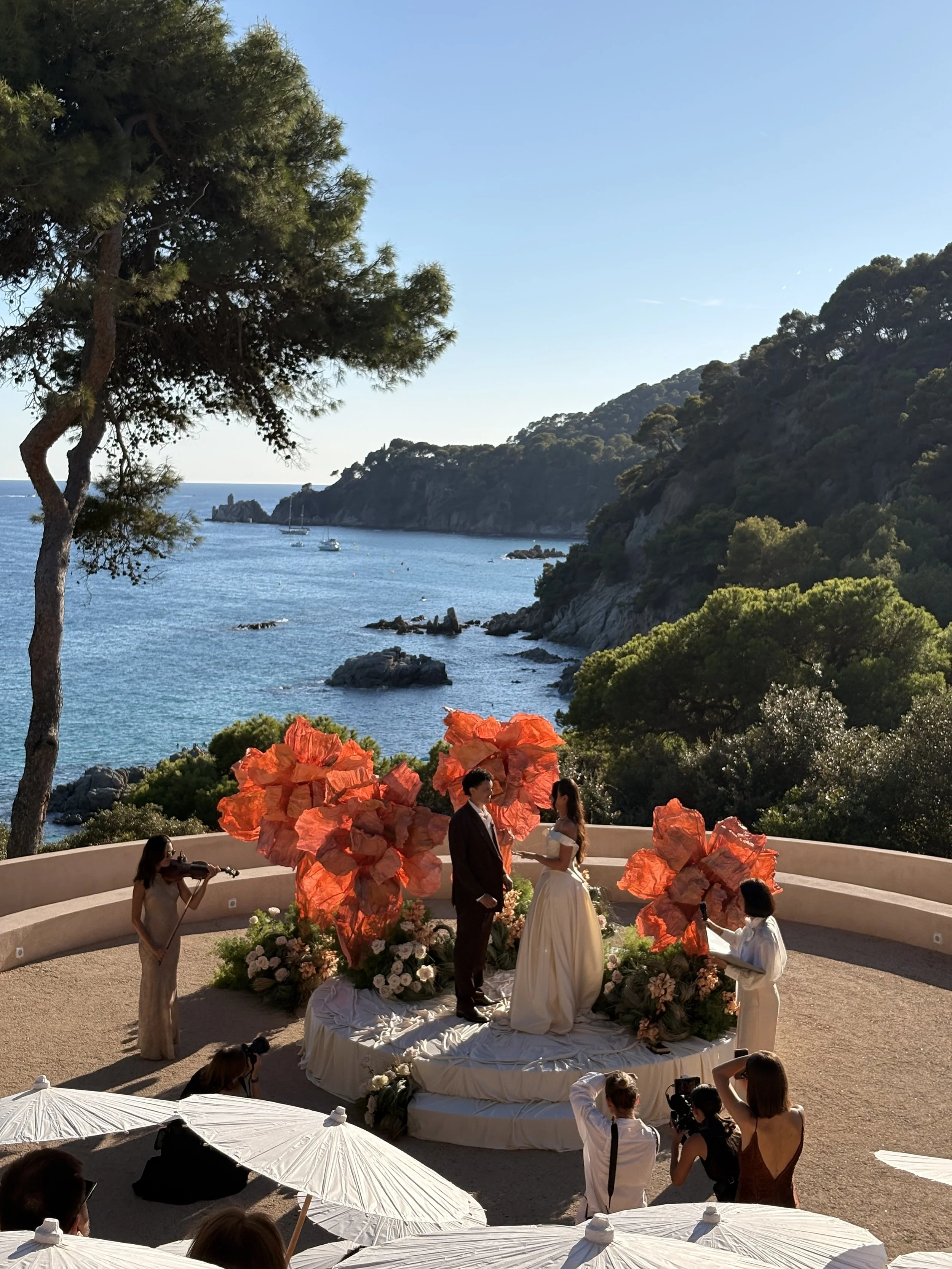 A wedding ceremony taking place outdoors near a scenic coastline with boats and a rocky hill in the background. The couple is standing at the altar, surrounded by floral arrangements and orange paper decorations. Bridesmaids and officiants are present, with guests seated under white umbrellas.