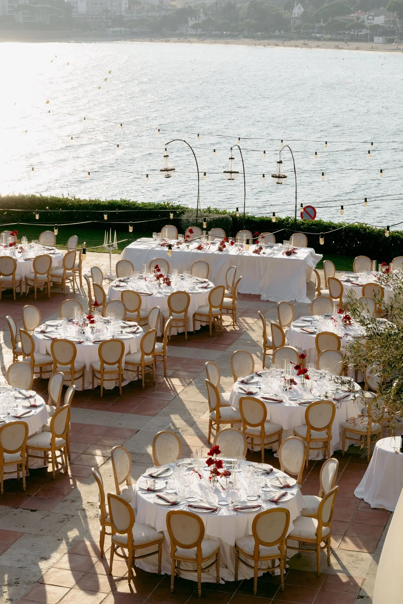 Outdoor dining setup with round tables covered in white tablecloths, decorated with red flowers, set with glassware, plates, and silverware, overlooking a beach and water with string lights hanging overhead in the daytime.