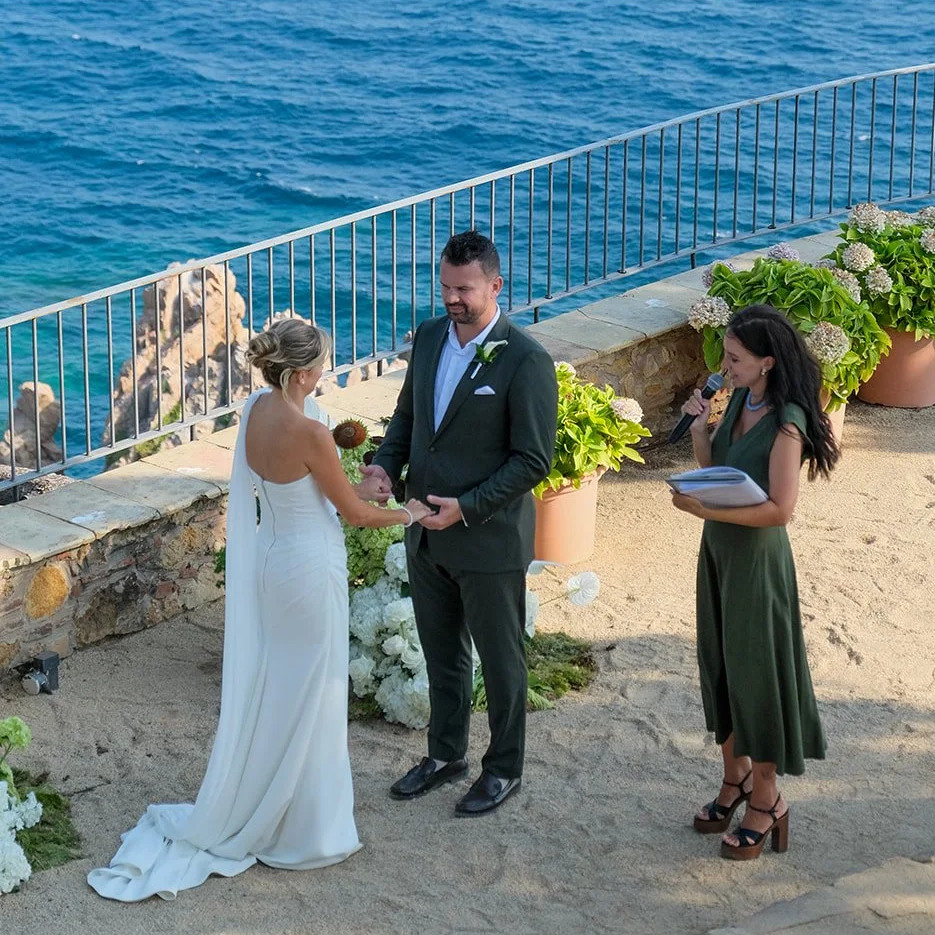 A couple getting married outdoors by the ocean, with an officiant reading and a woman holding a microphone, surrounded by potted plants and flowers.