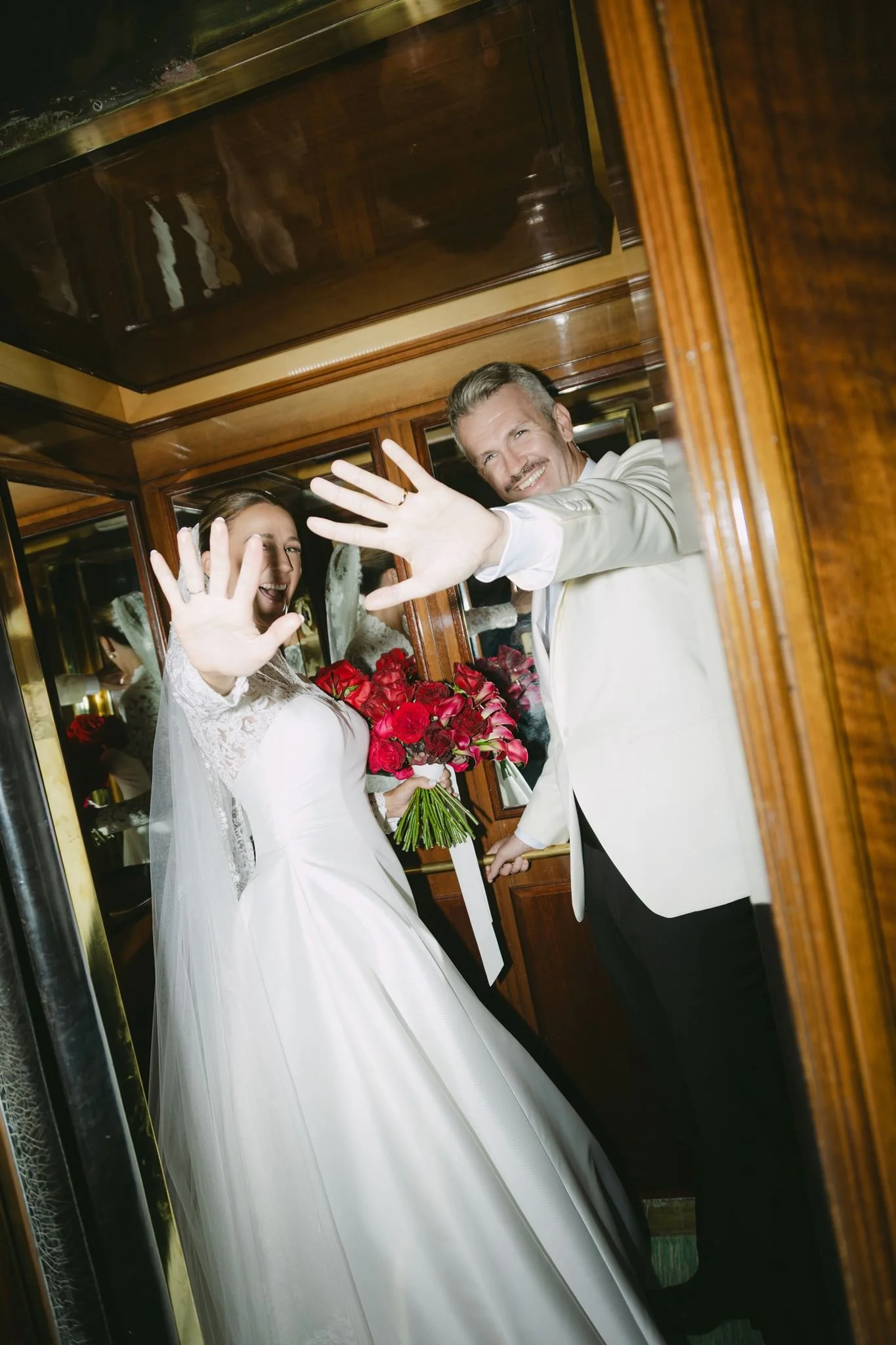 A bride and groom waving through a glass window, with the bride holding a bouquet of red roses, both smiling.