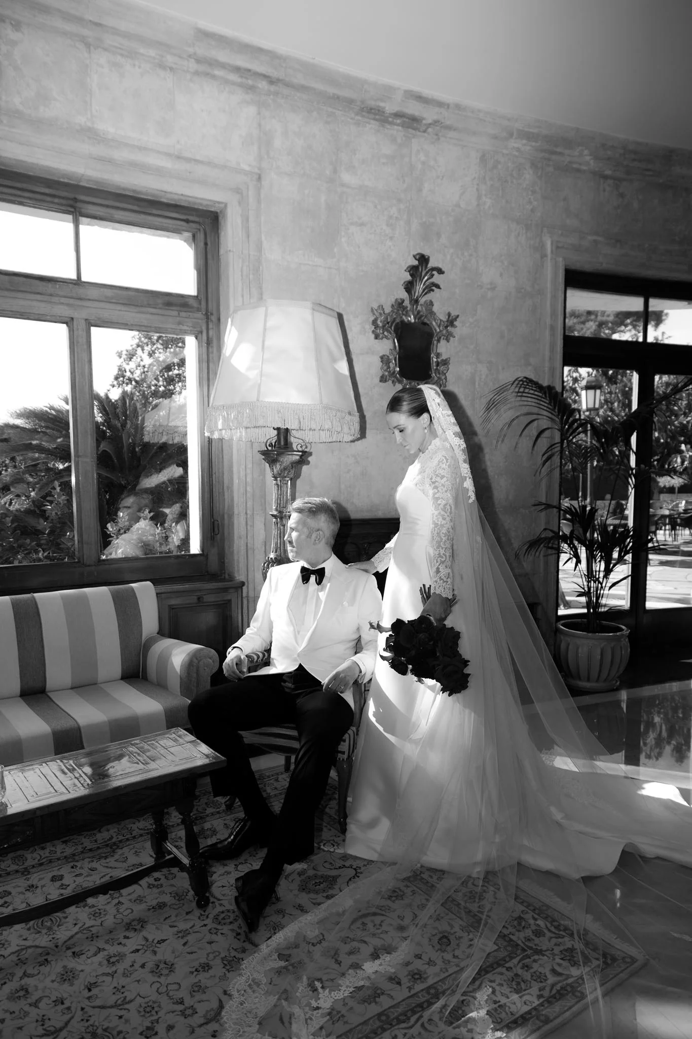 Black and white photo of a bride and groom in a room with large windows, decorated with antique furniture, plants, and an ornate mirror. The bride is standing holding a bouquet, and the groom is seated on a striped sofa.