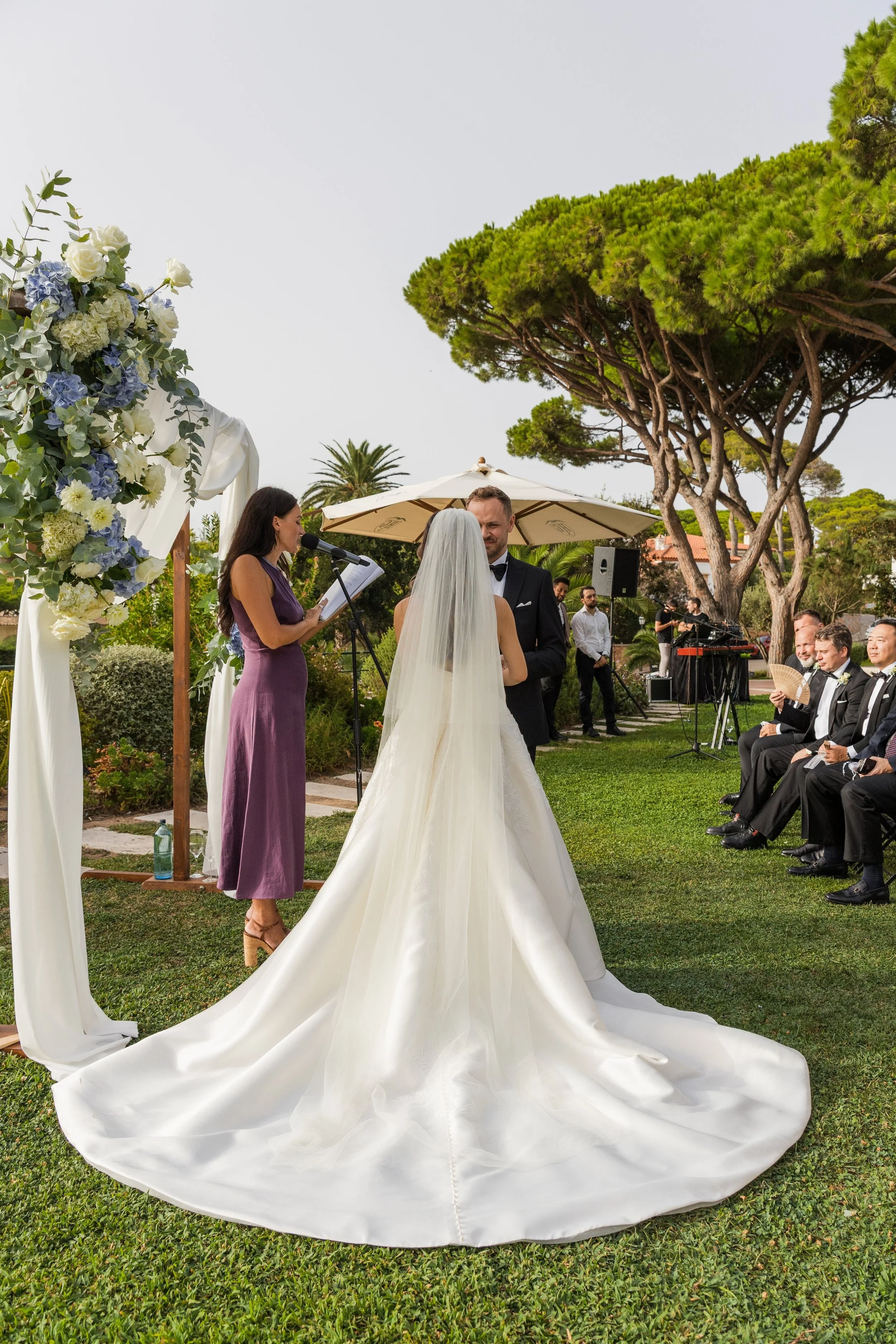 A wedding ceremony taking place outdoors with a couple facing each other, a woman officiating, and guests seated on the right. Decor includes floral arrangements and a white draped arch. Large trees and an umbrella are in the background.