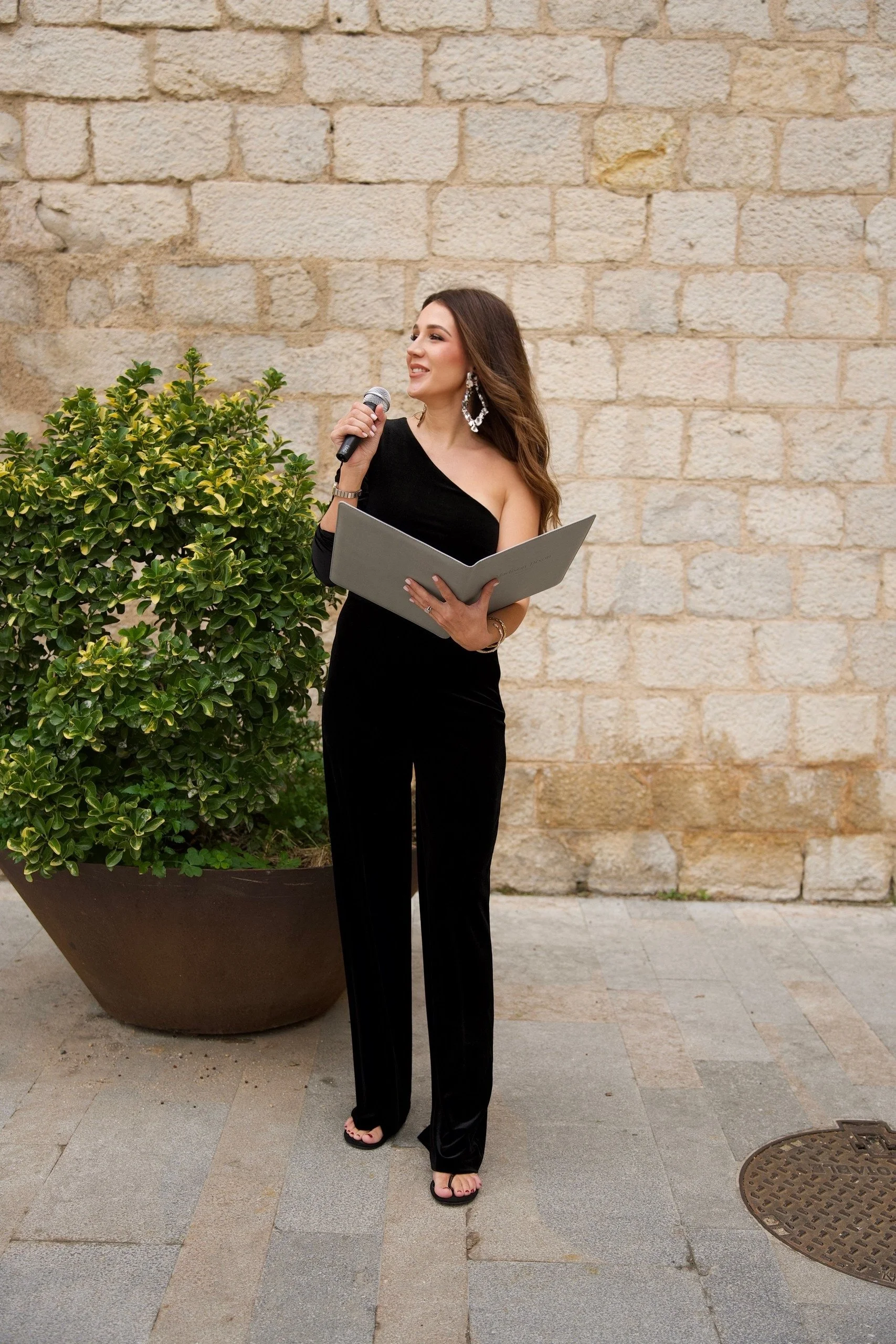 A woman in a black, one-shoulder gown standing outdoors in front of a beige brick wall, holding a microphone and a folder, next to a large potted green shrub.