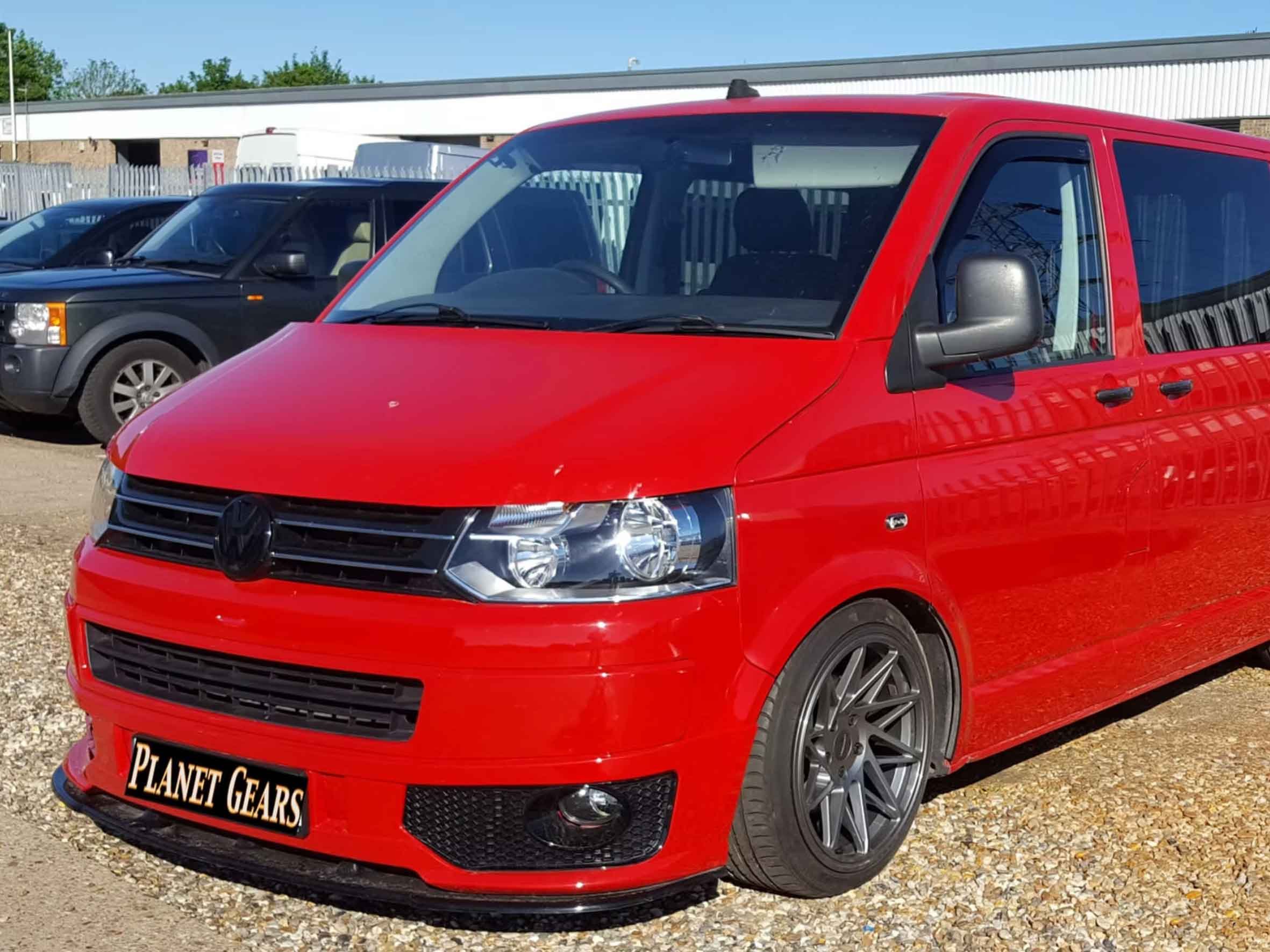 Red Volkswagen camper van parked outdoors with other vehicles in the background on a gravel lot.