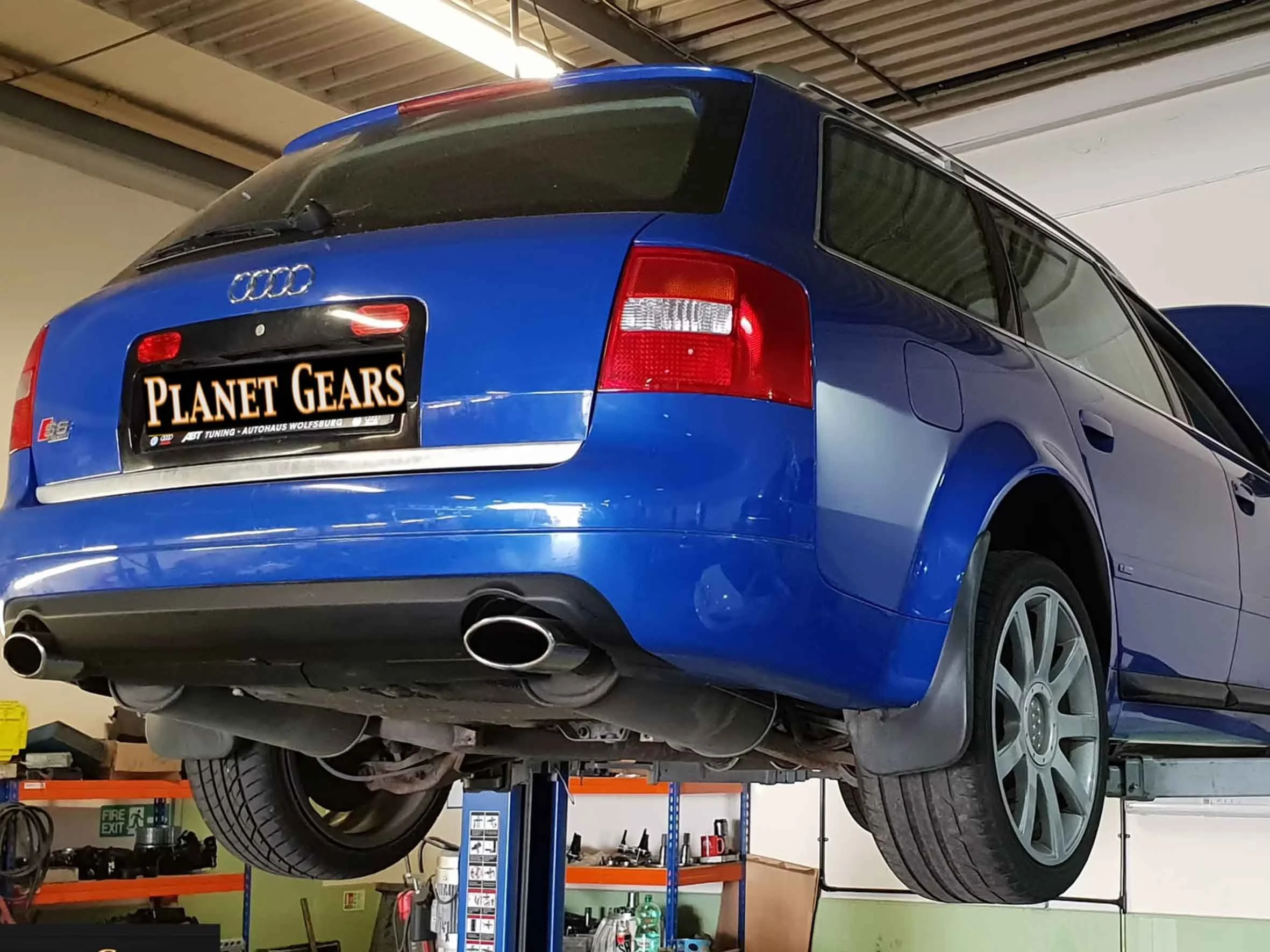 A blue Audi station wagon inside a garage, elevated on a lift, showing the underside and rear tires, with a 'Planet Gears' sign on the license plate area.