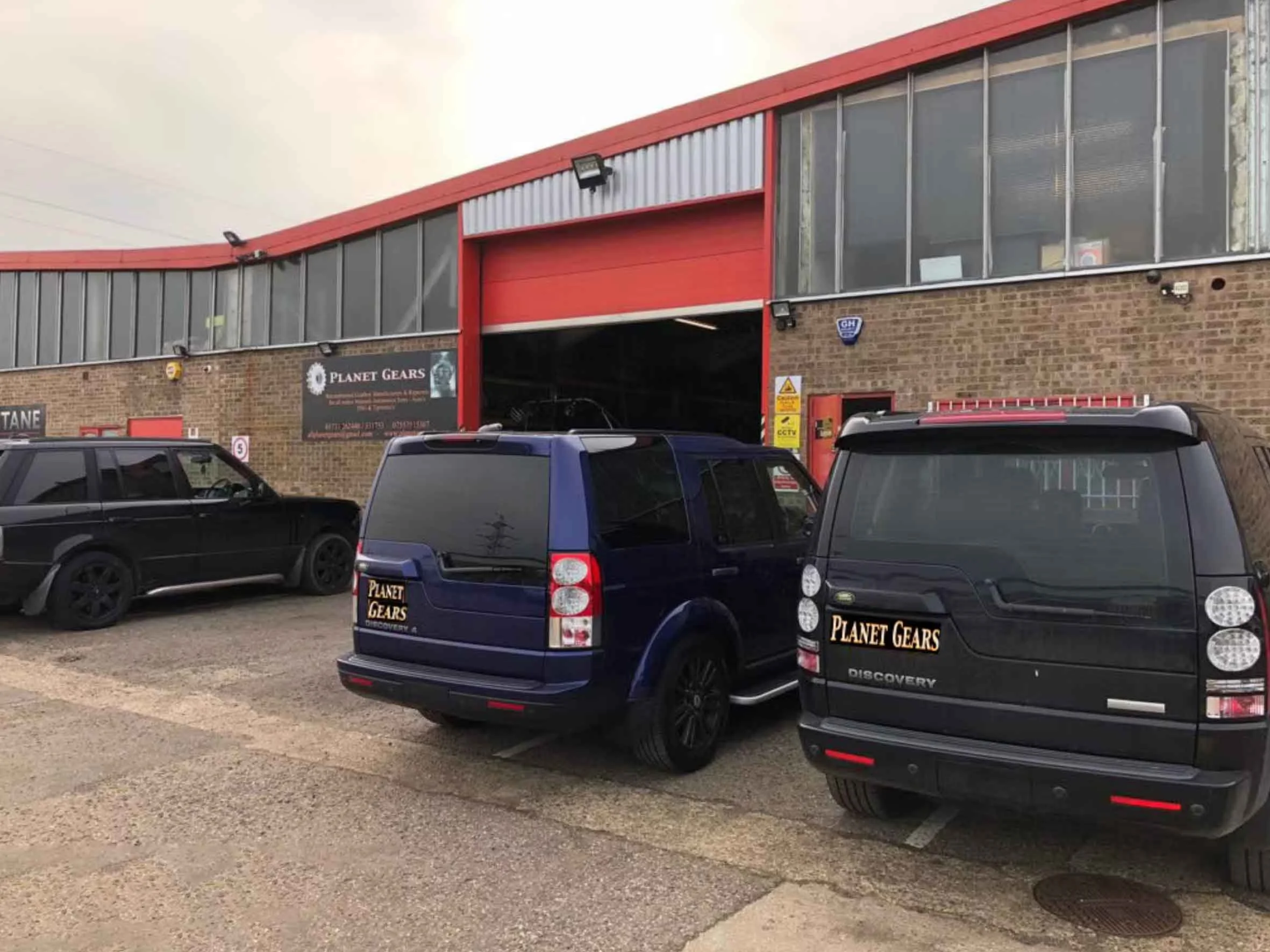 Parking lot outside a brick building with a red metal garage door, and vehicles with 'Planet Gears' stickers, including a black and a blue SUV, and a black car to the side.