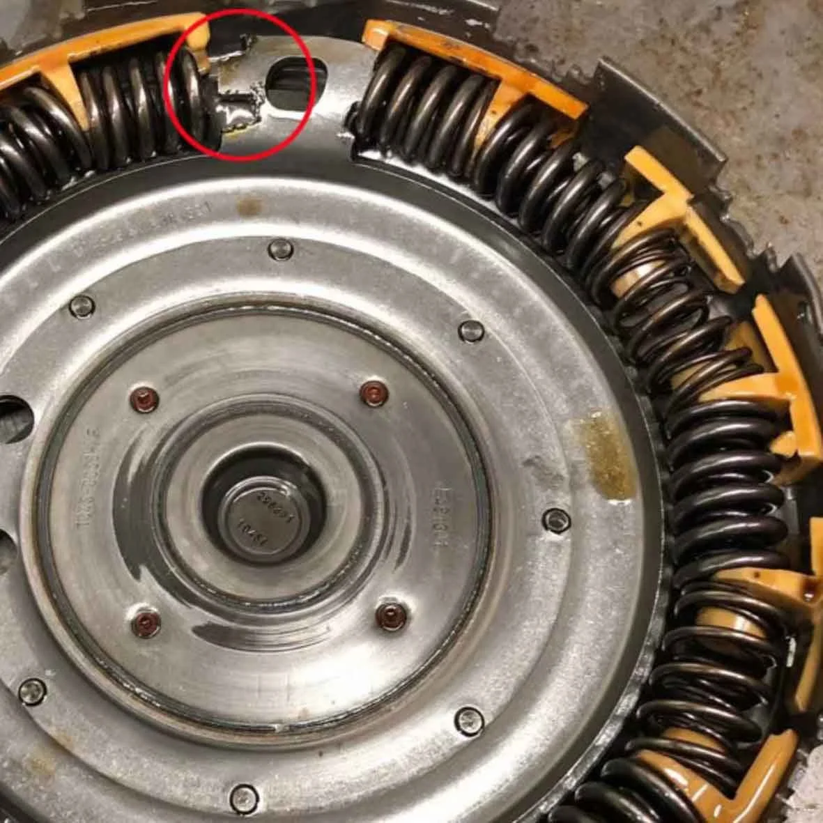 Close-up of a mechanical device with a circular metal component and a damaged coil spring circled in red.