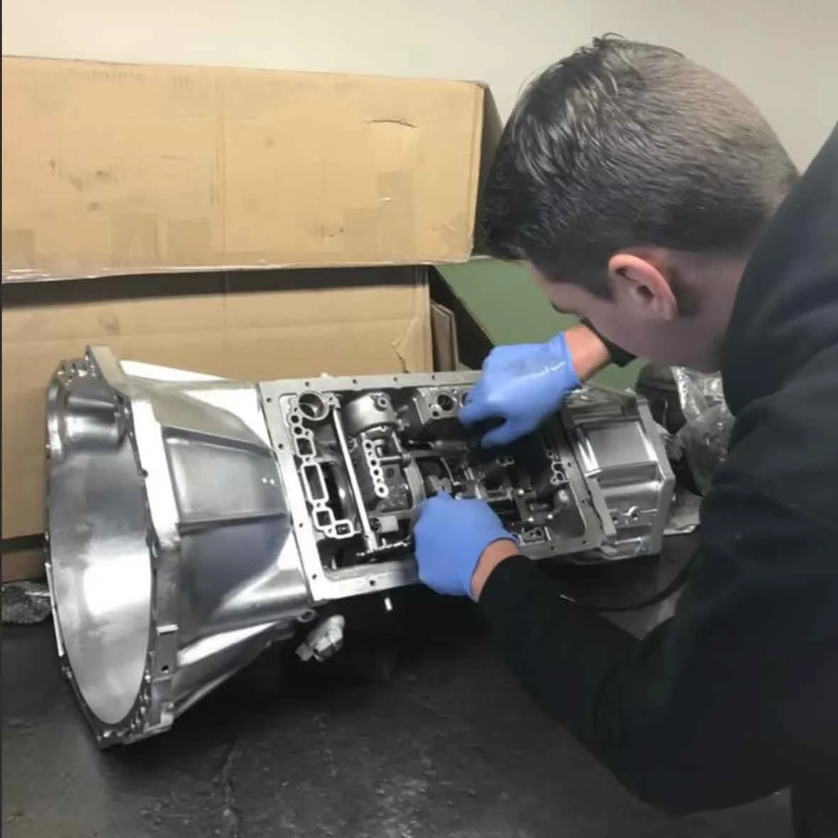 A man wearing blue gloves working on a metallic engine or mechanical component in a workshop.