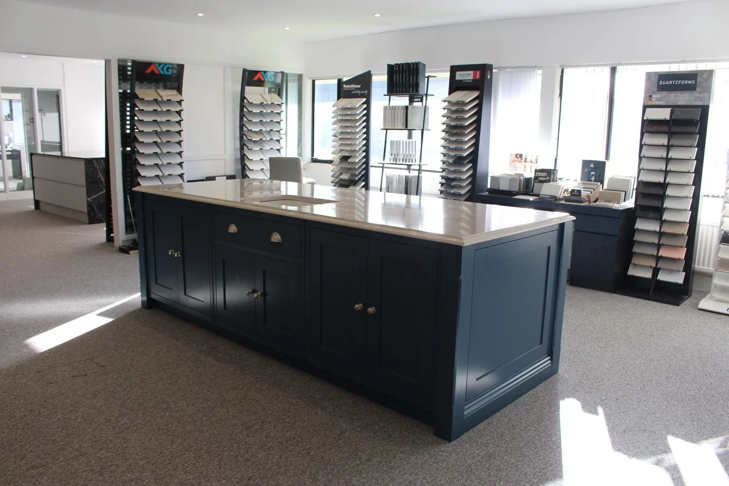Showroom with a large blue island counter, sample swatches and display racks of materials in a well-lit office or retail space.