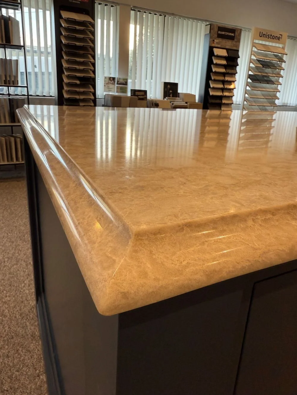 Close-up of a polished wooden countertop in a showroom, with display racks and sample materials in the background.