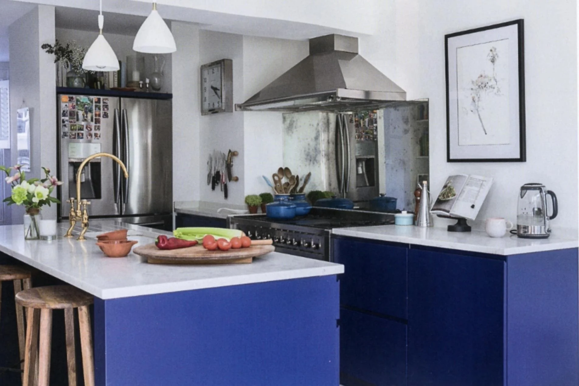 A modern kitchen with a white countertop, blue cabinets, and stainless steel appliances. There are fresh vegetables on a wooden cutting board and a vase with flowers on the counter. A picture frame and kitchen utensils are visible on the wall.