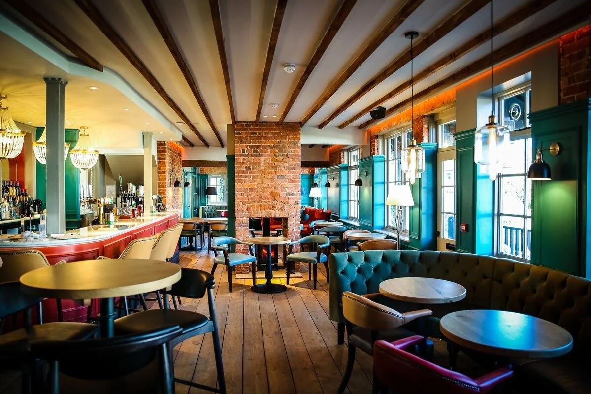Interior of a colorful bar and restaurant with wooden floors, exposed brick walls, and large windows with blue frames, decorated with hanging lights and lamps.