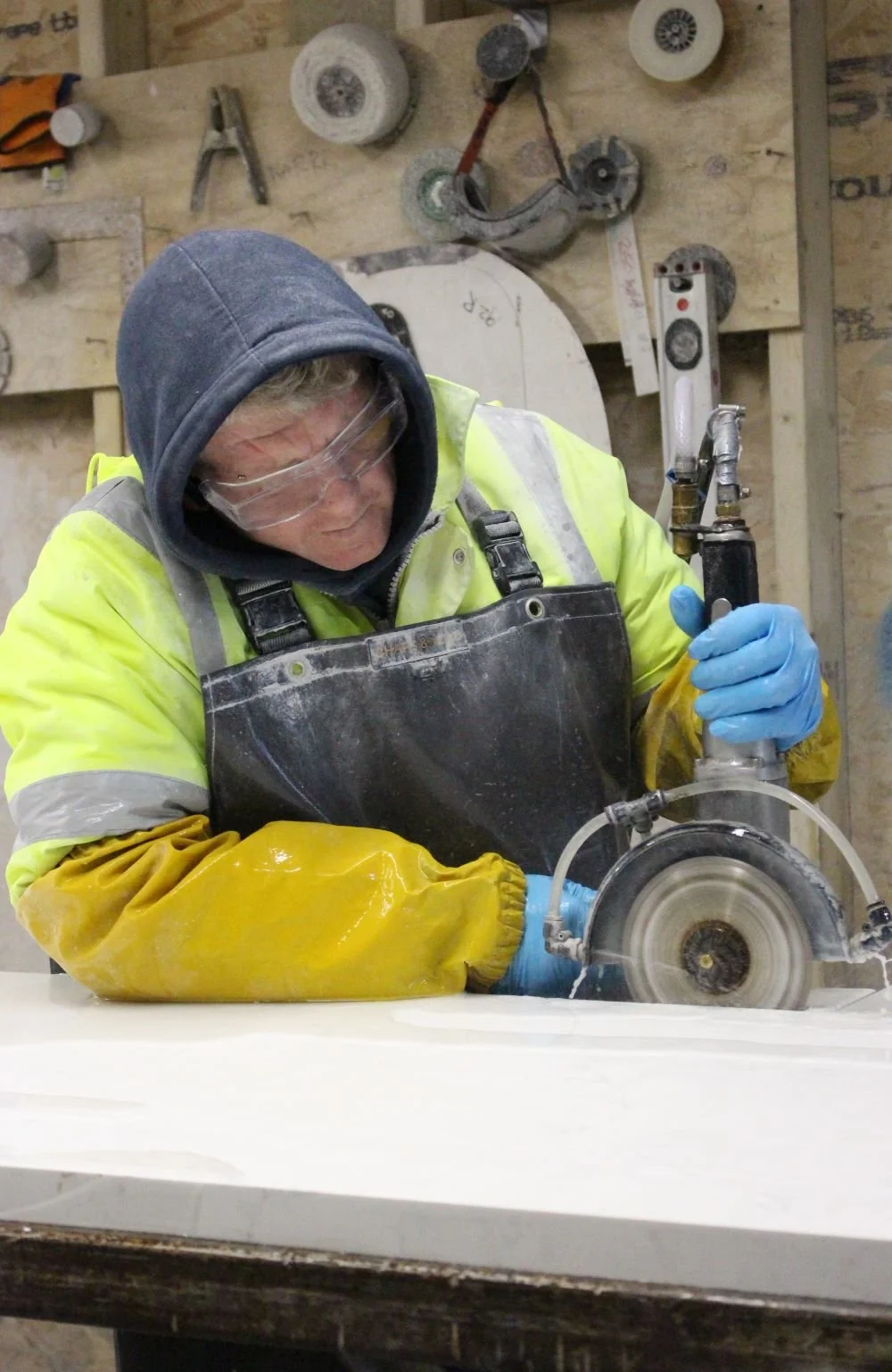 A person wearing safety glasses, a hoodie, a high-visibility jacket, and blue gloves operates a power tool to cut a large sheet of white material in a woodworking shop.
