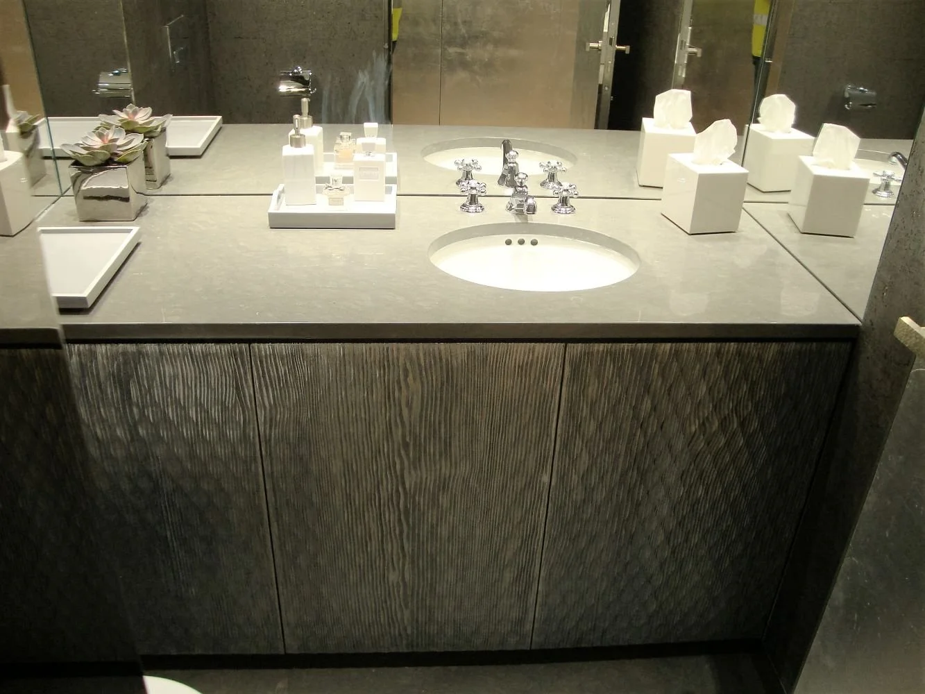 Bathroom sink vanity with a small round sink, silver faucet, and various toiletries and tissue boxes on the grey countertop, with a mirror reflecting the scene.