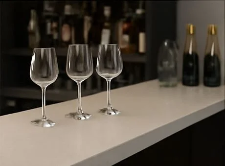 Three empty wine glasses on a white bar counter with bottles in the background.