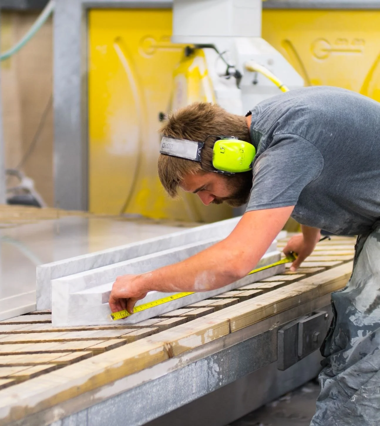 A man wearing safety headphones and work clothes is measuring a piece of material on a workbench in a workshop.