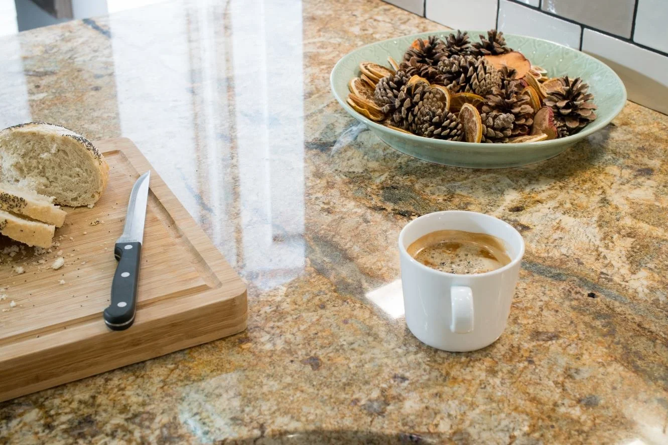 A kitchen countertop with a partially sliced loaf of bread on a wooden cutting board, a knife, a bowl filled with pinecones and dried citrus slices, and a white mug of coffee with crema.