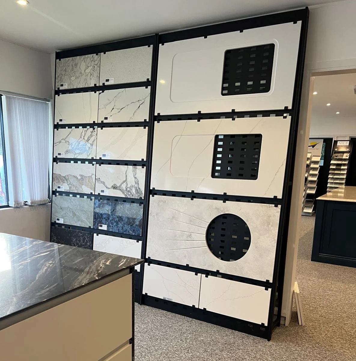 Display of various large marble and stone tile samples in a showroom, arranged on black-framed panels.