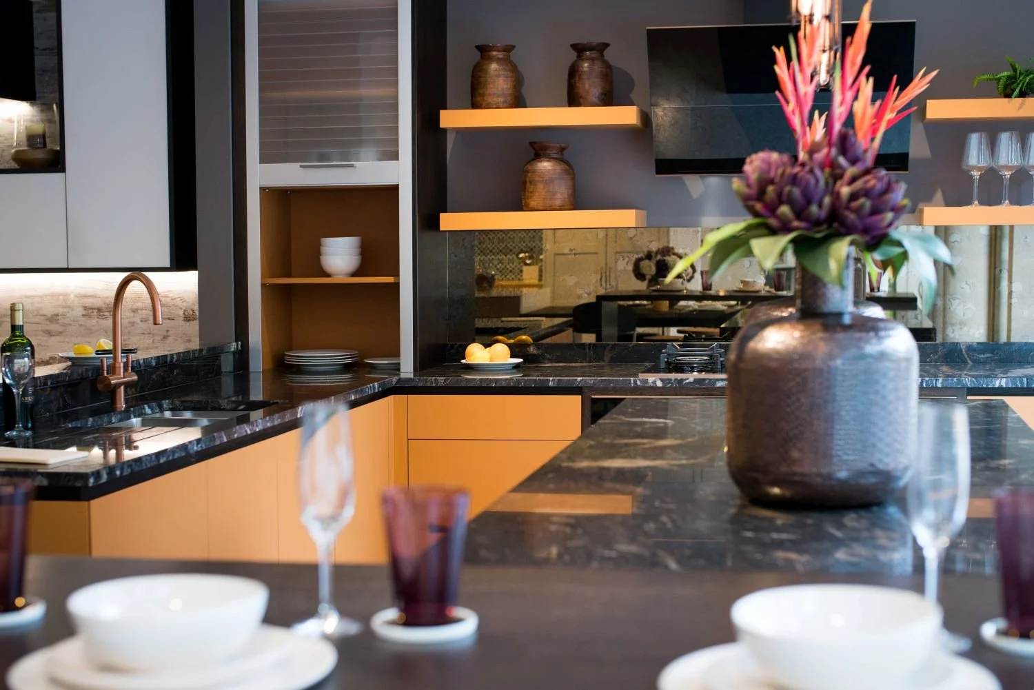 Modern kitchen with black marble countertops, orange cabinets, and open shelves displaying brown vases and white bowls. A large brown vase with purple and pink flowers is on the foreground island, with a dining table set with wine glasses and bowls.