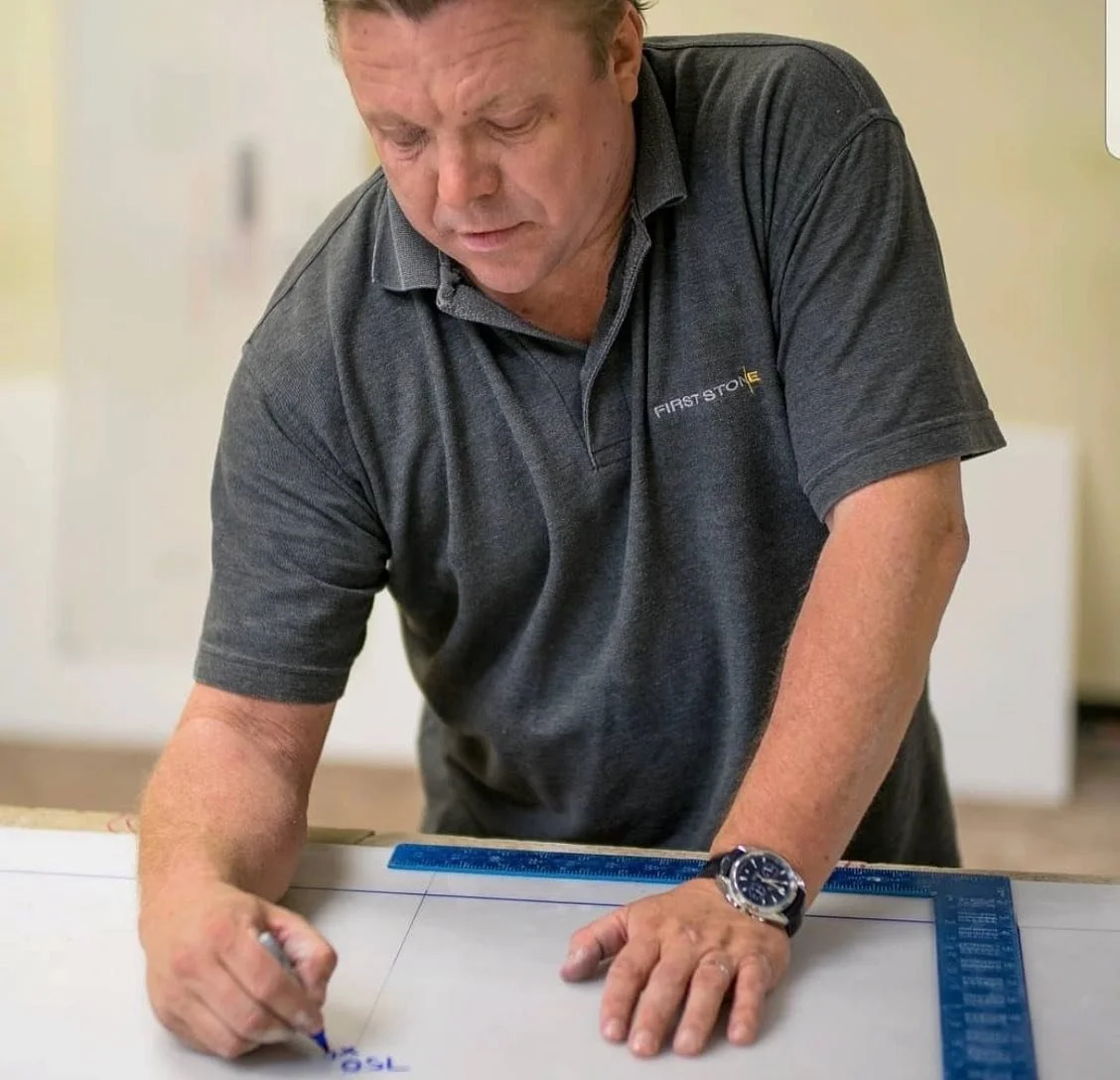 A man in a dark gray polo shirt marked with the text 'First Solo' is drawing or writing on a large whiteboard or paper with a blue marker. He is wearing a black watch and appears focused on his work.
