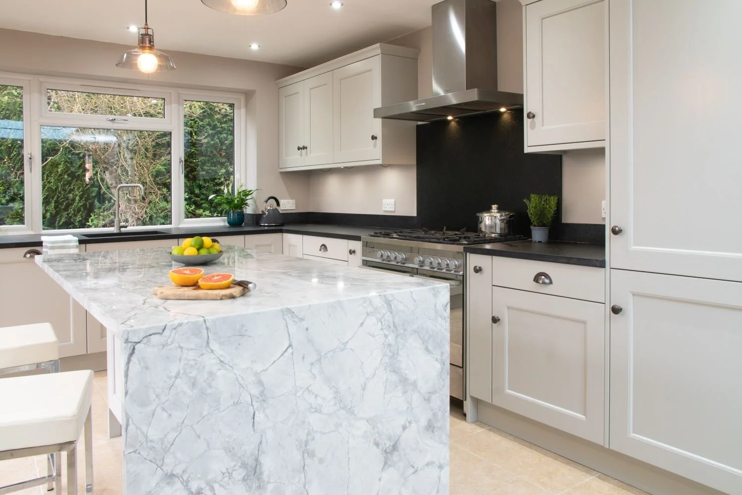 Modern kitchen with white cabinets, black countertops, and a marble island. A window overlooks greenery, with a bowl of lemons and grapefruit on the island. There is a stovetop, oven, and a range hood, with a plant and a kettle on the counters.