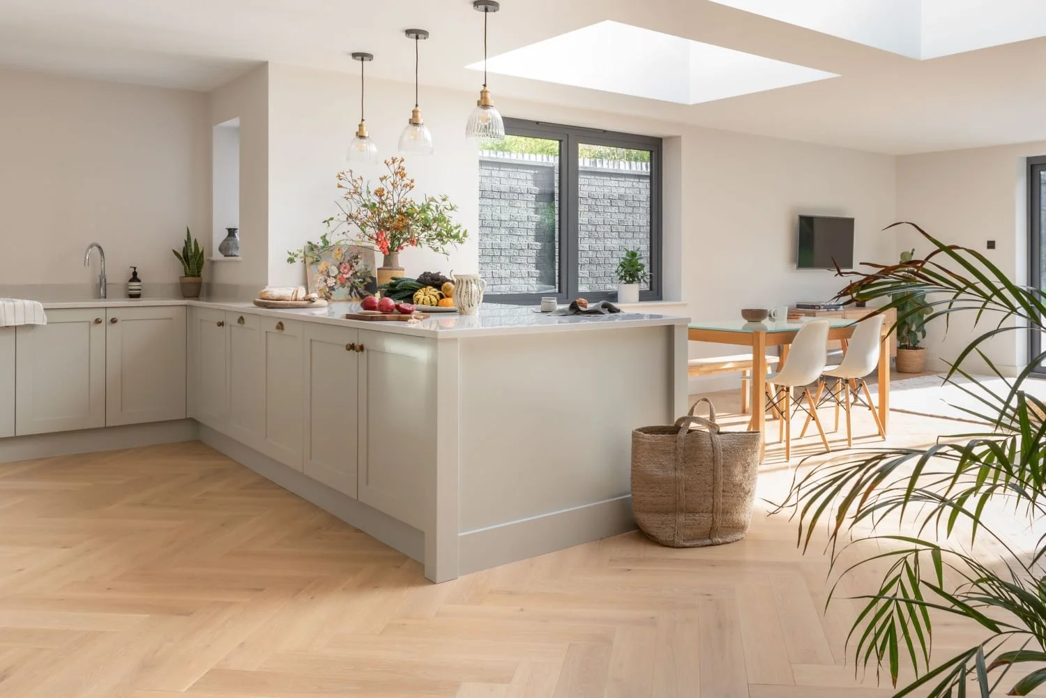 Bright, modern kitchen and dining area with white cabinetry, a kitchen island with various fruits and flowers, hanging pendant lights, a window, a wooden dining table with four white chairs, a wall-mounted TV, and several indoor plants.