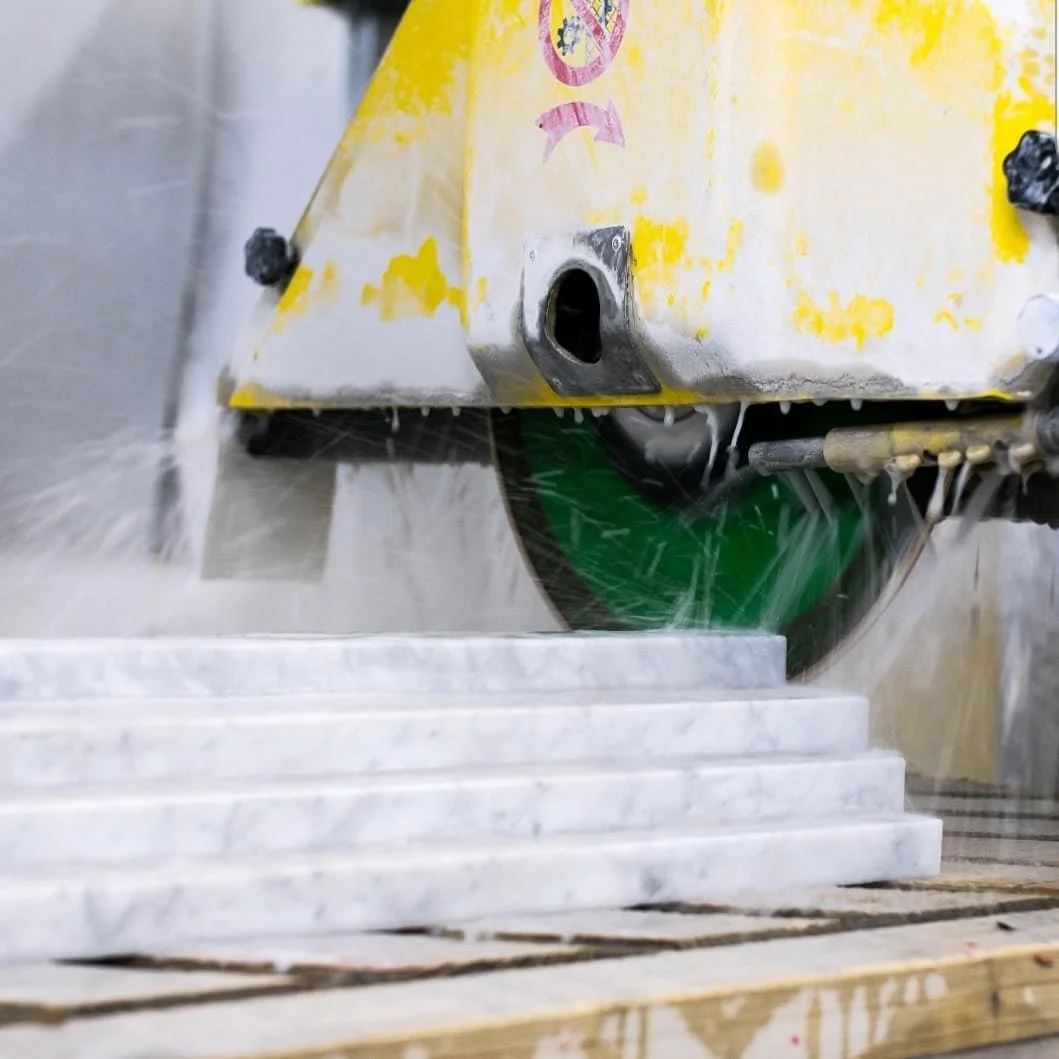 A marble cutting machine with a green blade cutting white marble slabs, producing water spray and marble dust.