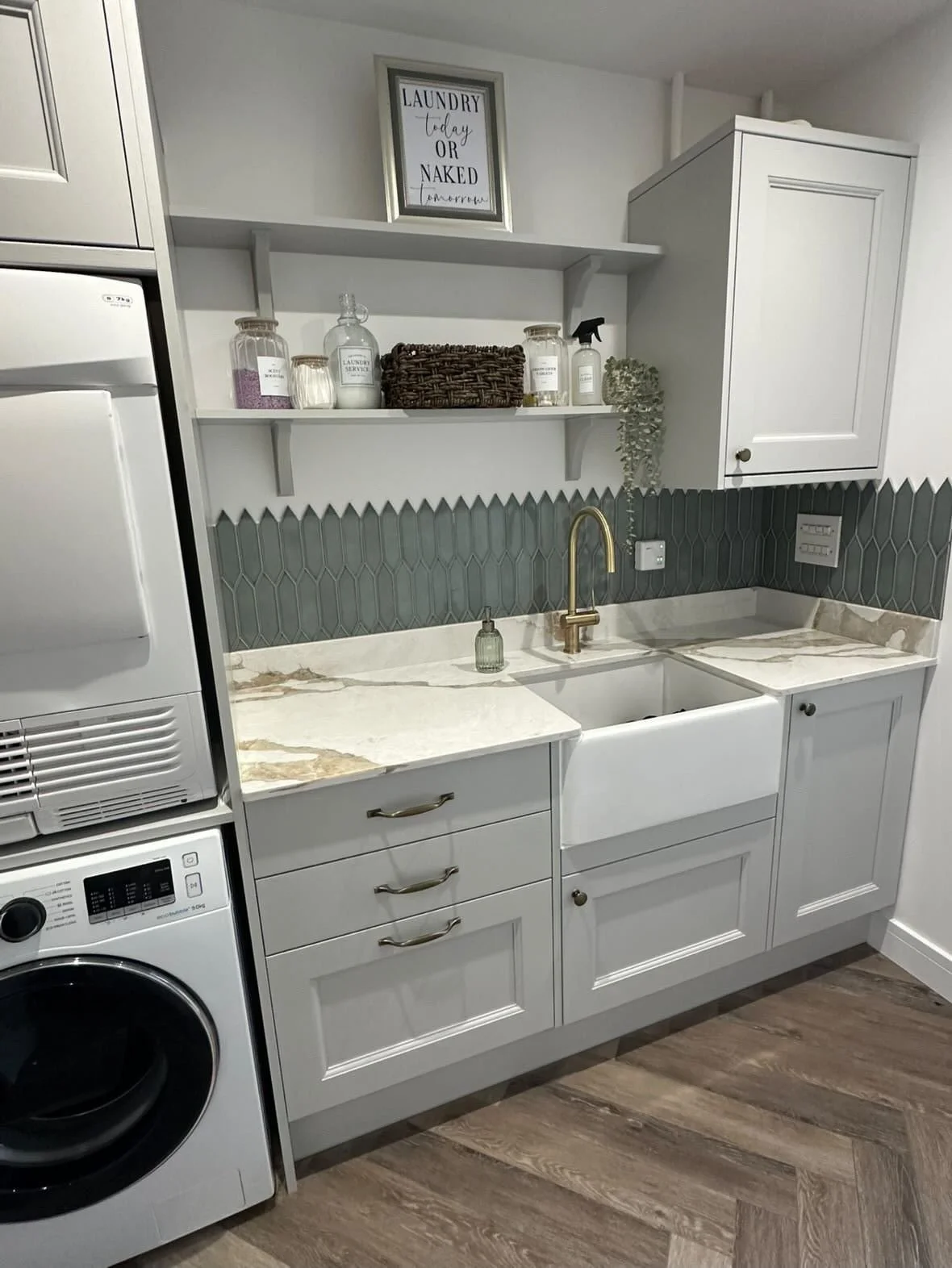 Small laundry room with white cabinetry, a marble countertop, green tile backsplash, farmhouse sink, gold faucet, open shelving with jars and decor, washing machine, framed sign that reads 'Laundry today or naked tomorrow', wood flooring.