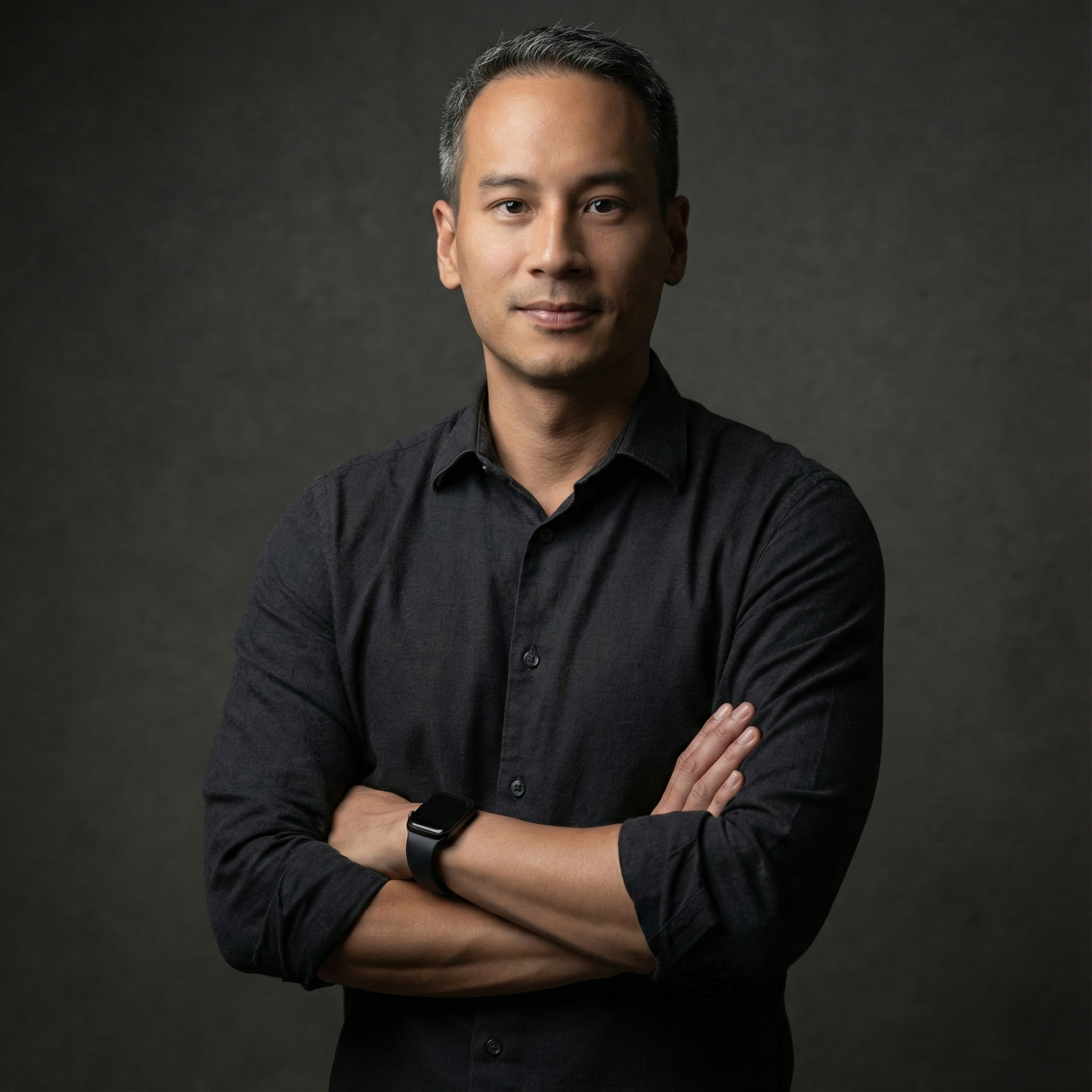 Portrait of a man with crossed arms, wearing a black shirt and a smartwatch, standing against a dark grey background.