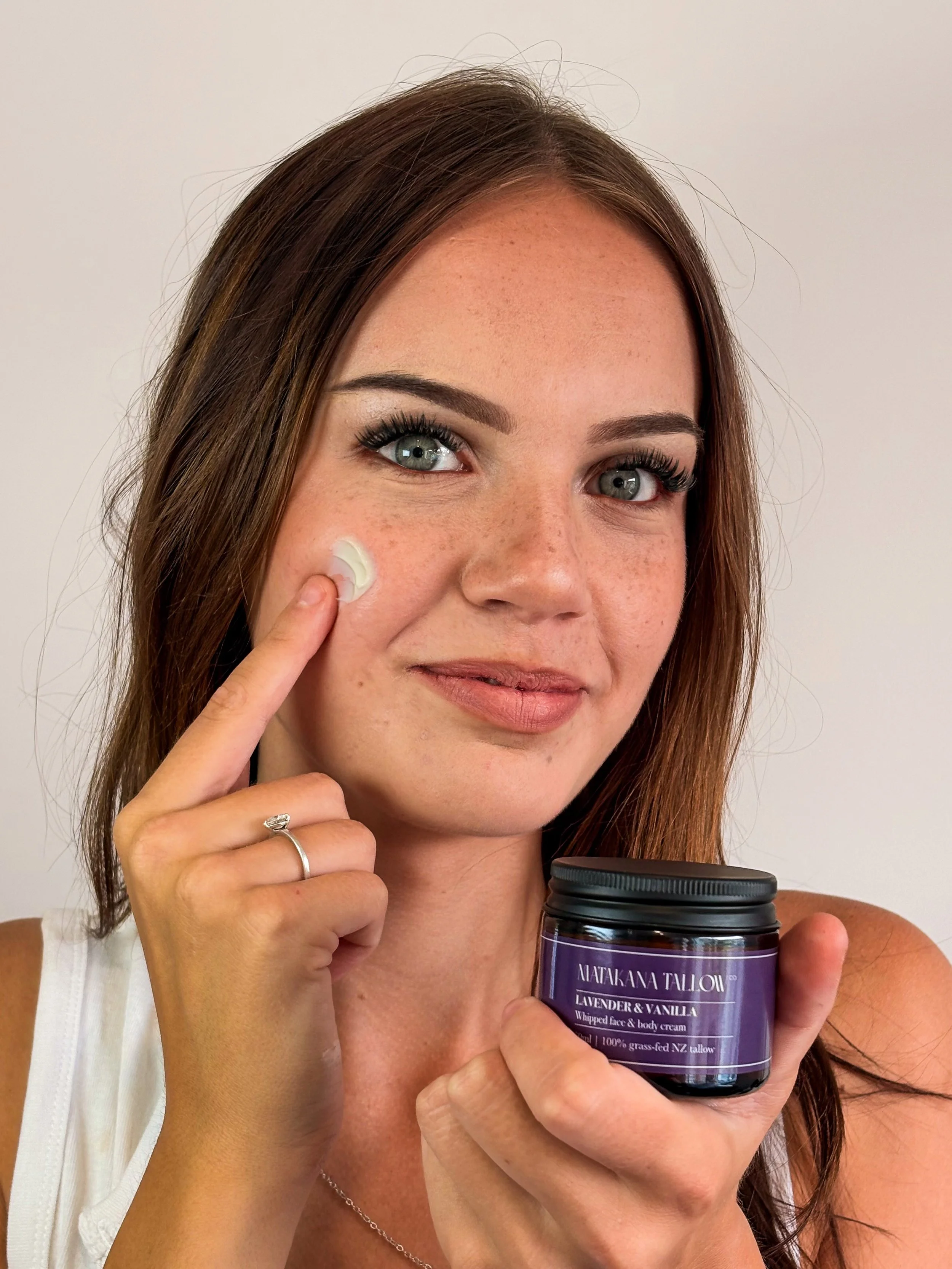 Woman with red hair and blue eyes holding a jar of Matakana tallow co lavender and vanilla whipped face and body cream, applying cream to her face.