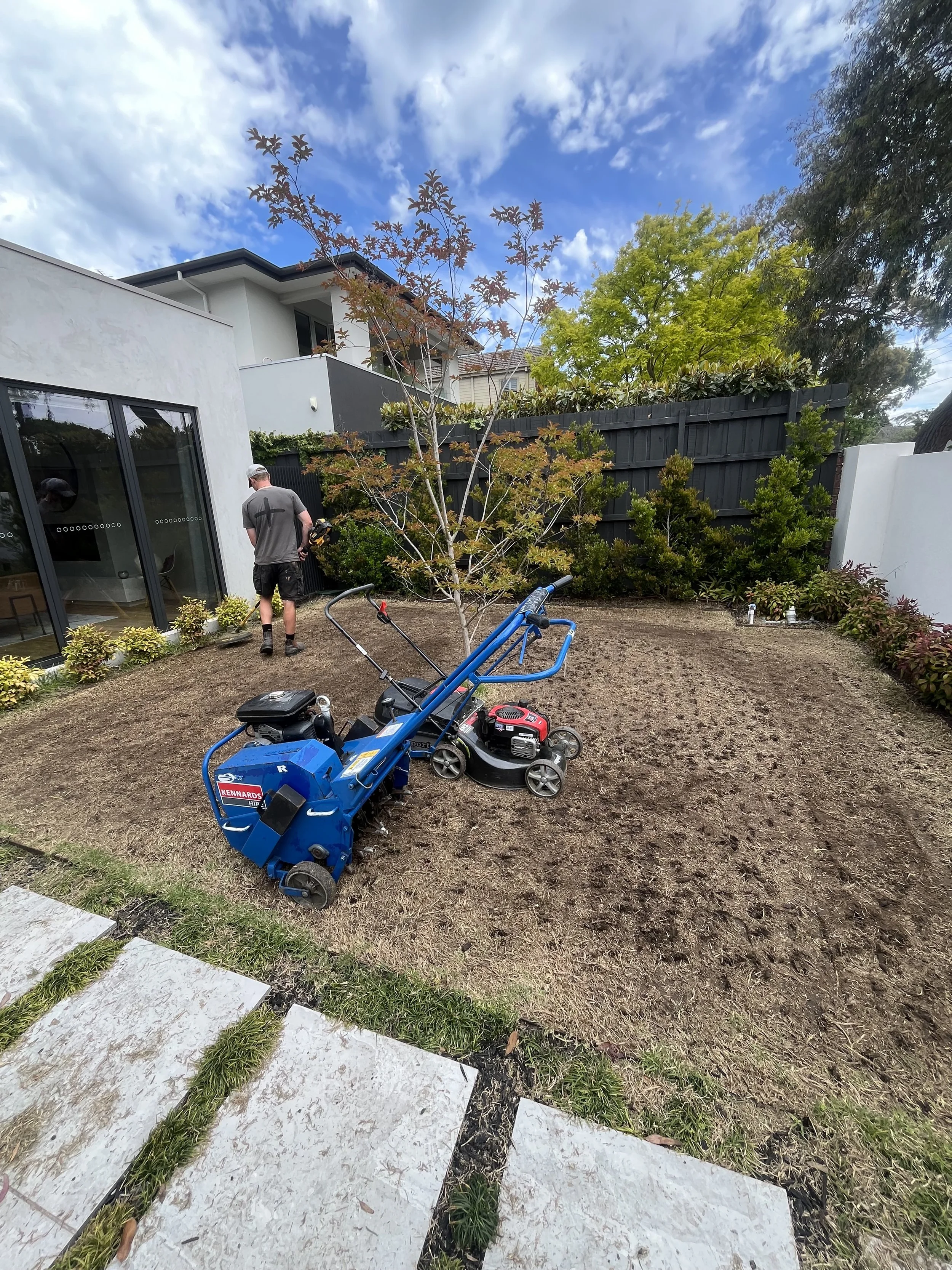 Backyard with a person doing yard work near a black fence, a small tree, bushes, a lawn mower, and a leaf blower. Modern house with floor-to-ceiling windows and cloudy blue sky.