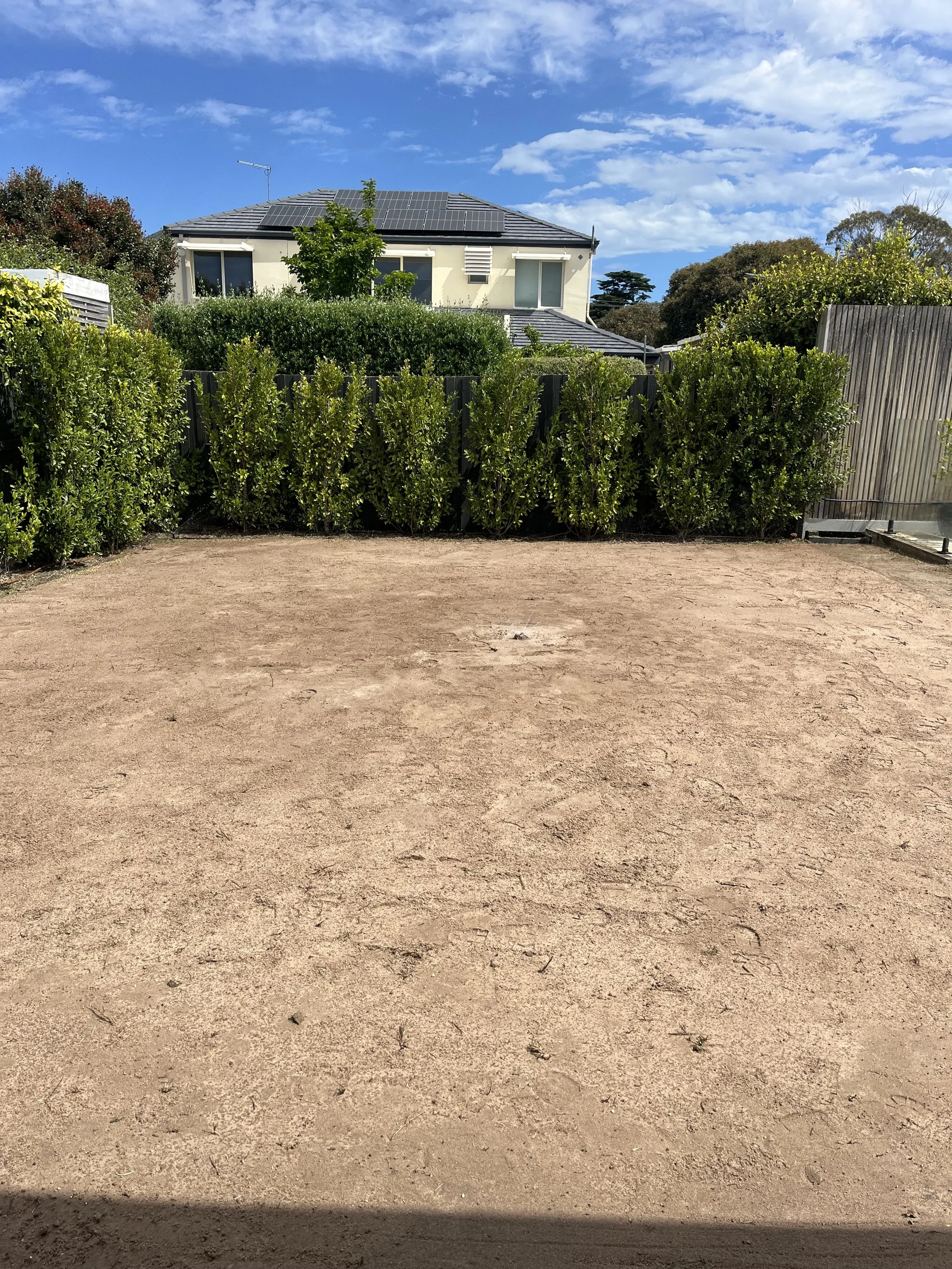 Empty lot with dirt ground, green shrubbery in the background, and a house with solar panels on the roof, under a partly cloudy sky.