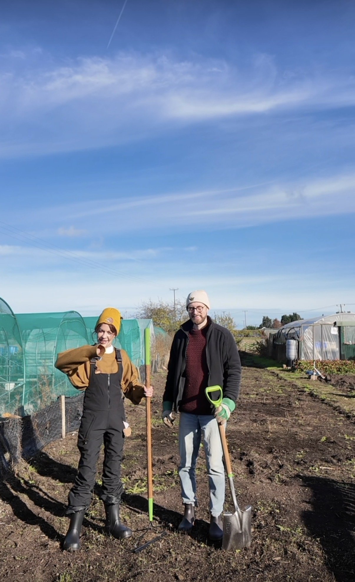 Two people standing in a garden on a bright, sunny day. They are wearing yard work clothes and holding gardening tools, with greenhouses and a clear blue sky in the background.