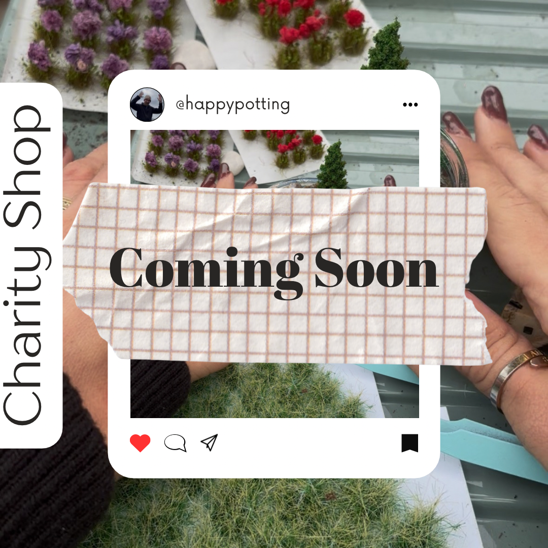 A cardboard sign with a grid pattern and the words 'Coming Soon' is held up by a person with painted fingernails. The background includes small flower plants and work surface.