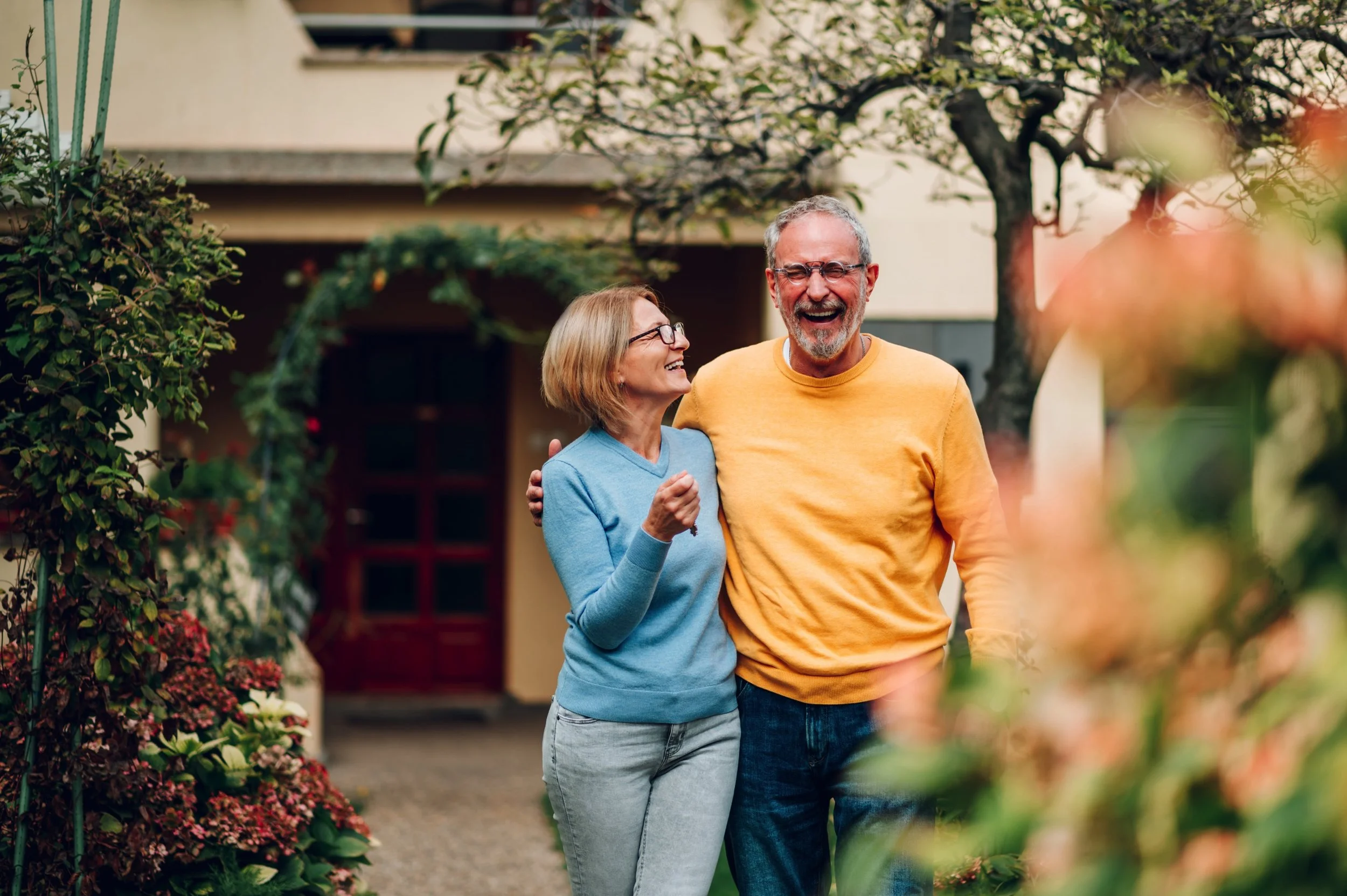 An elderly couple, a woman with glasses and a man with a beard and glasses, are walking outdoors, smiling and looking happy, with trees and a house in the background.