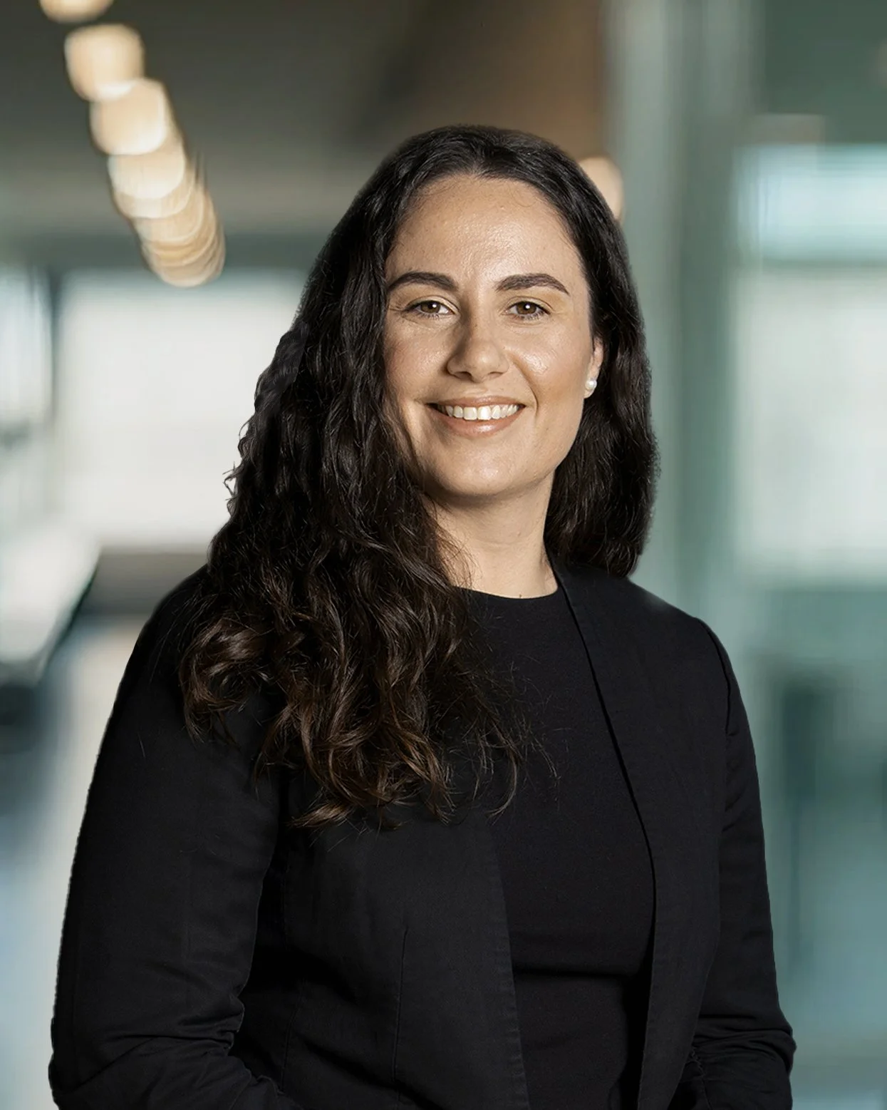 A professional woman with long curly dark hair and a bright smile, wearing a black blazer and pearl earrings, standing in a modern office corridor with blurred lights and windows in the background.