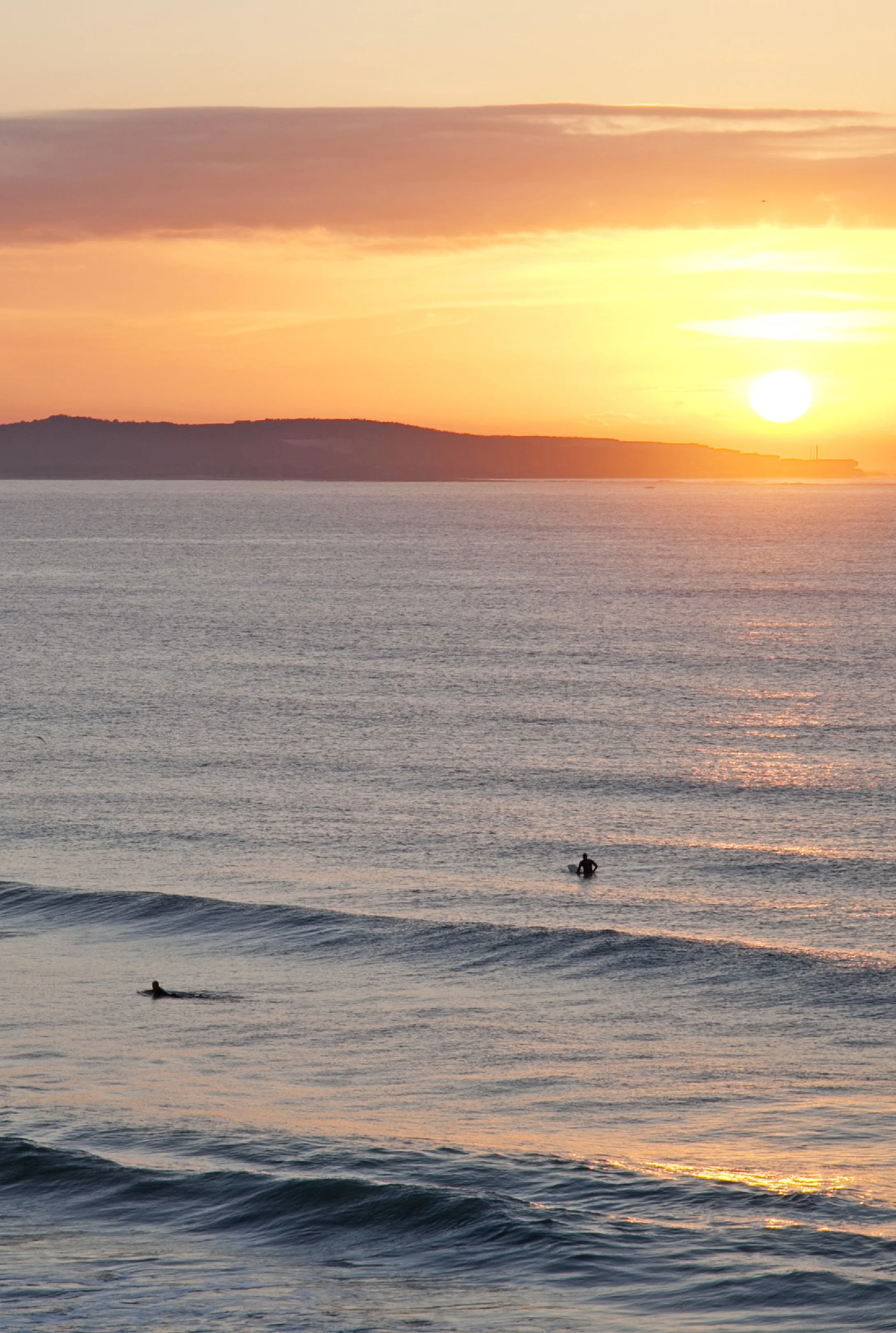 Sunset over the ocean with a person surfing and a swimmer in the water. The sky is orange and pink with clouds, and land is visible in the distance.
