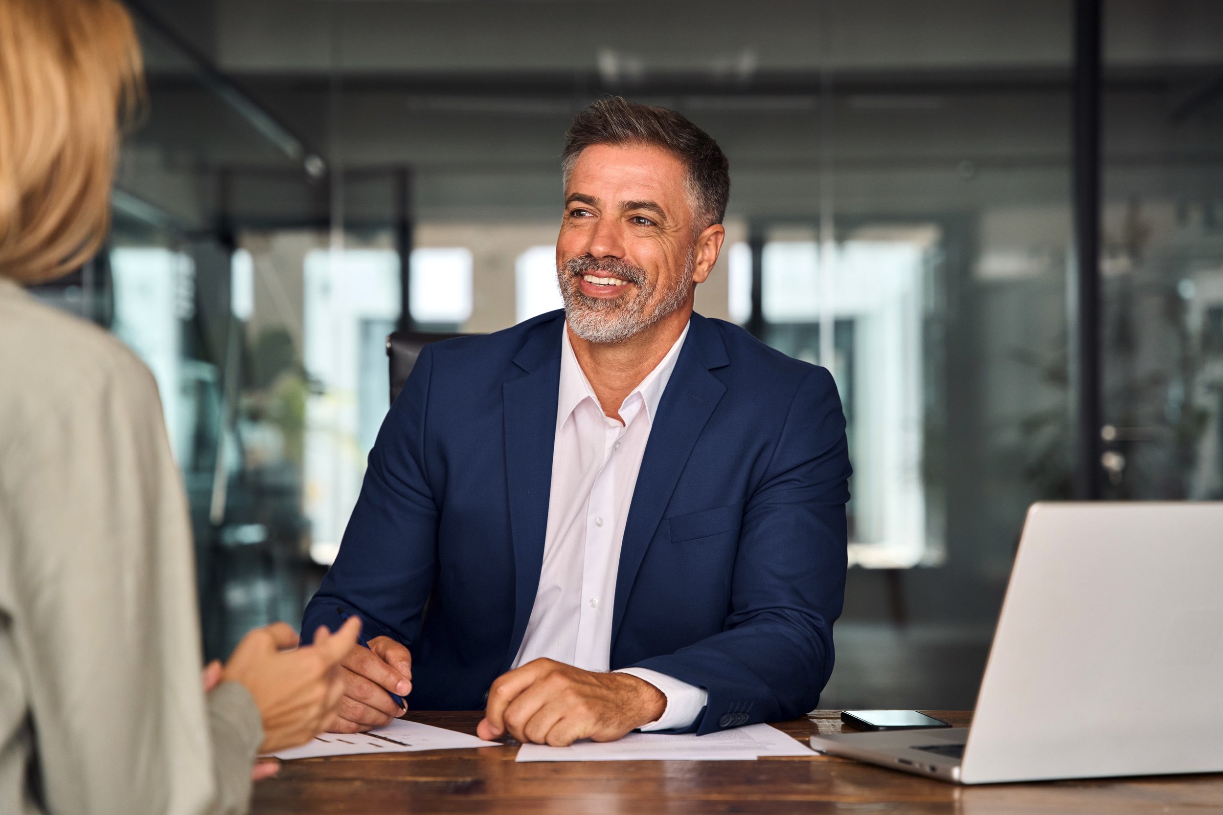 A middle-aged man in a blue suit and white shirt smiling while talking with a woman in a beige blazer during a meeting in a modern office.