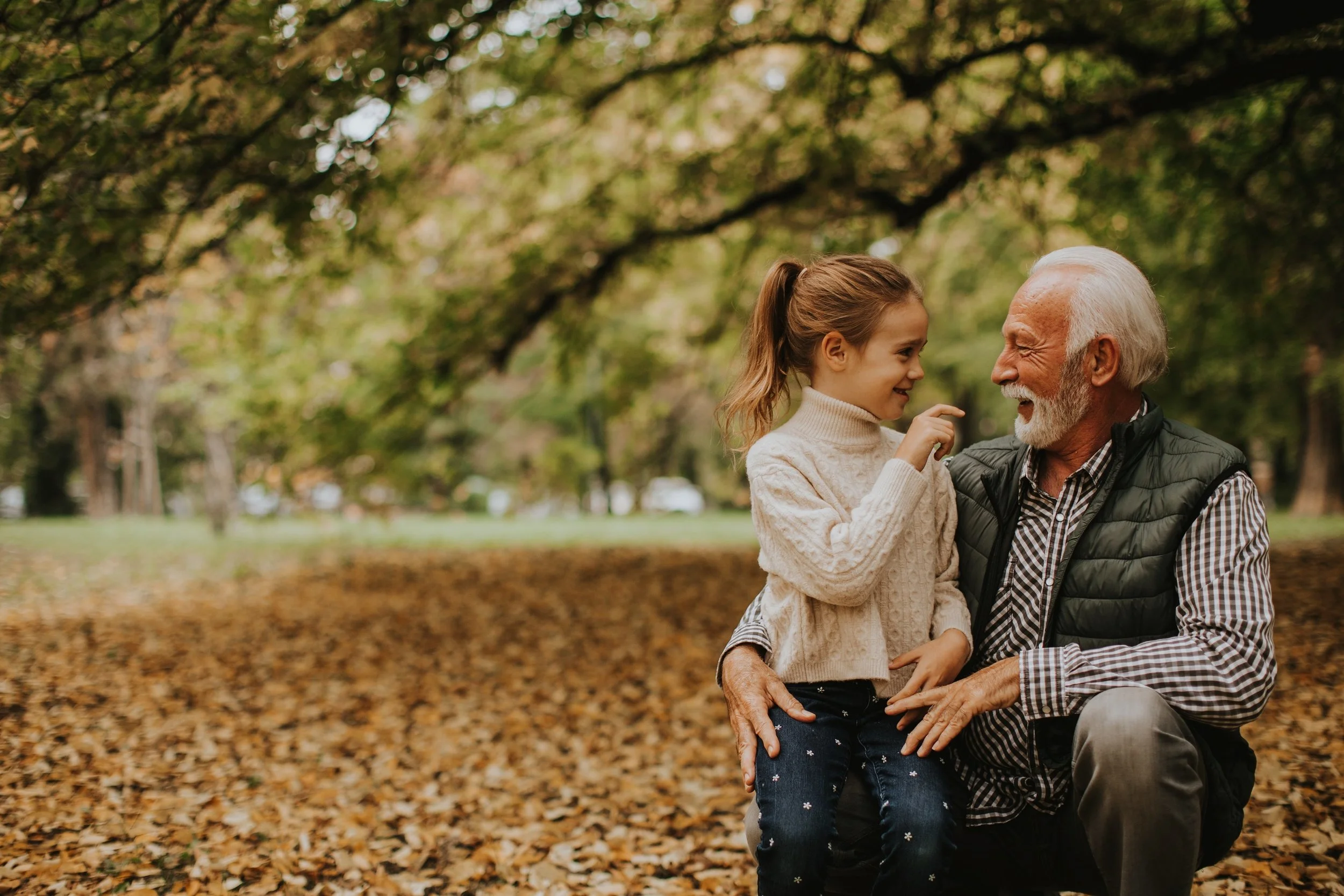 An elderly man and young girl enjoying a moment together on a fall day in a park, with leaves on the ground and trees in the background.