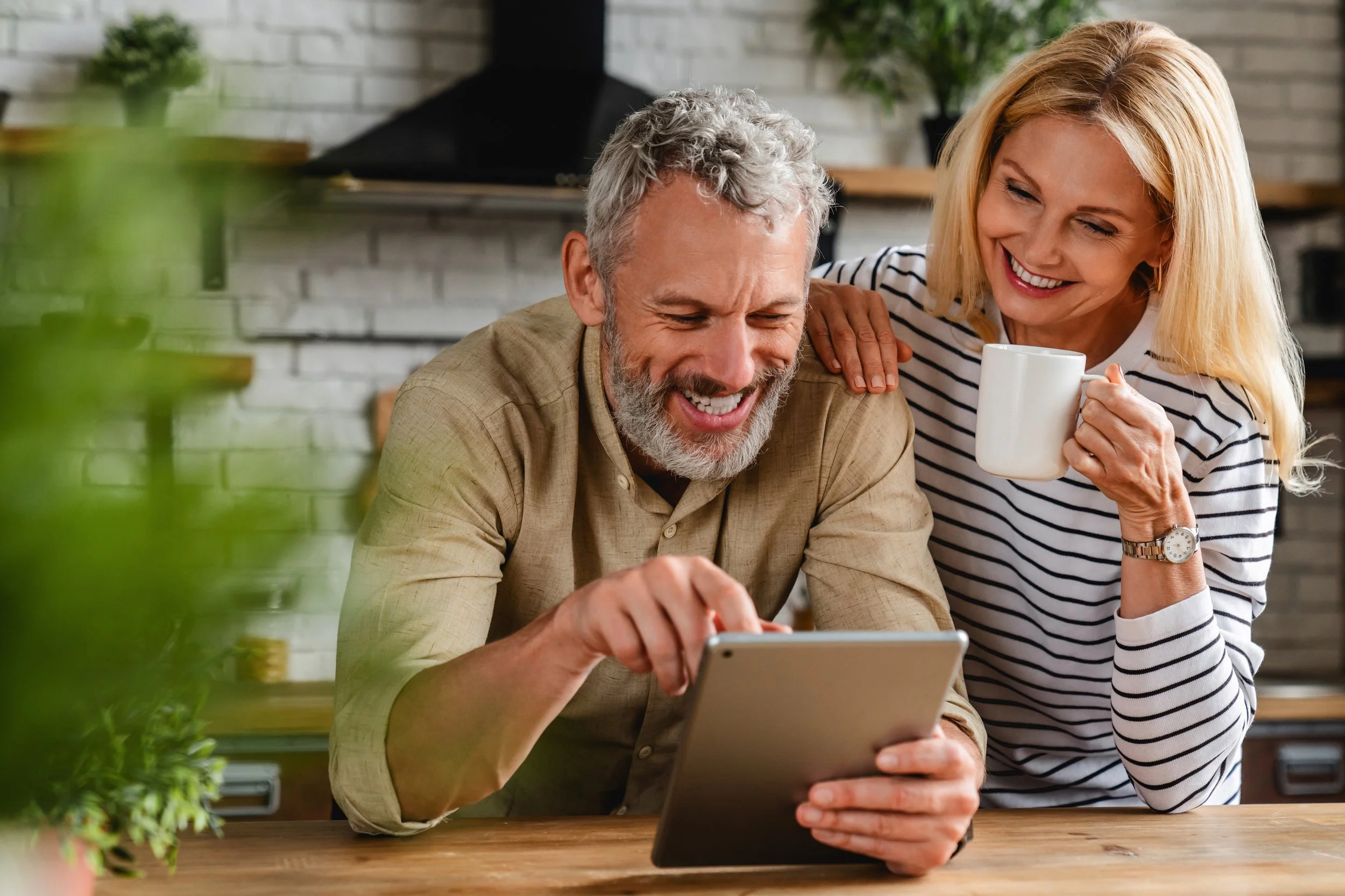 An older man and woman smiling while looking at a tablet, with the woman holding a mug and resting her hand on the man's shoulder in a cozy kitchen.